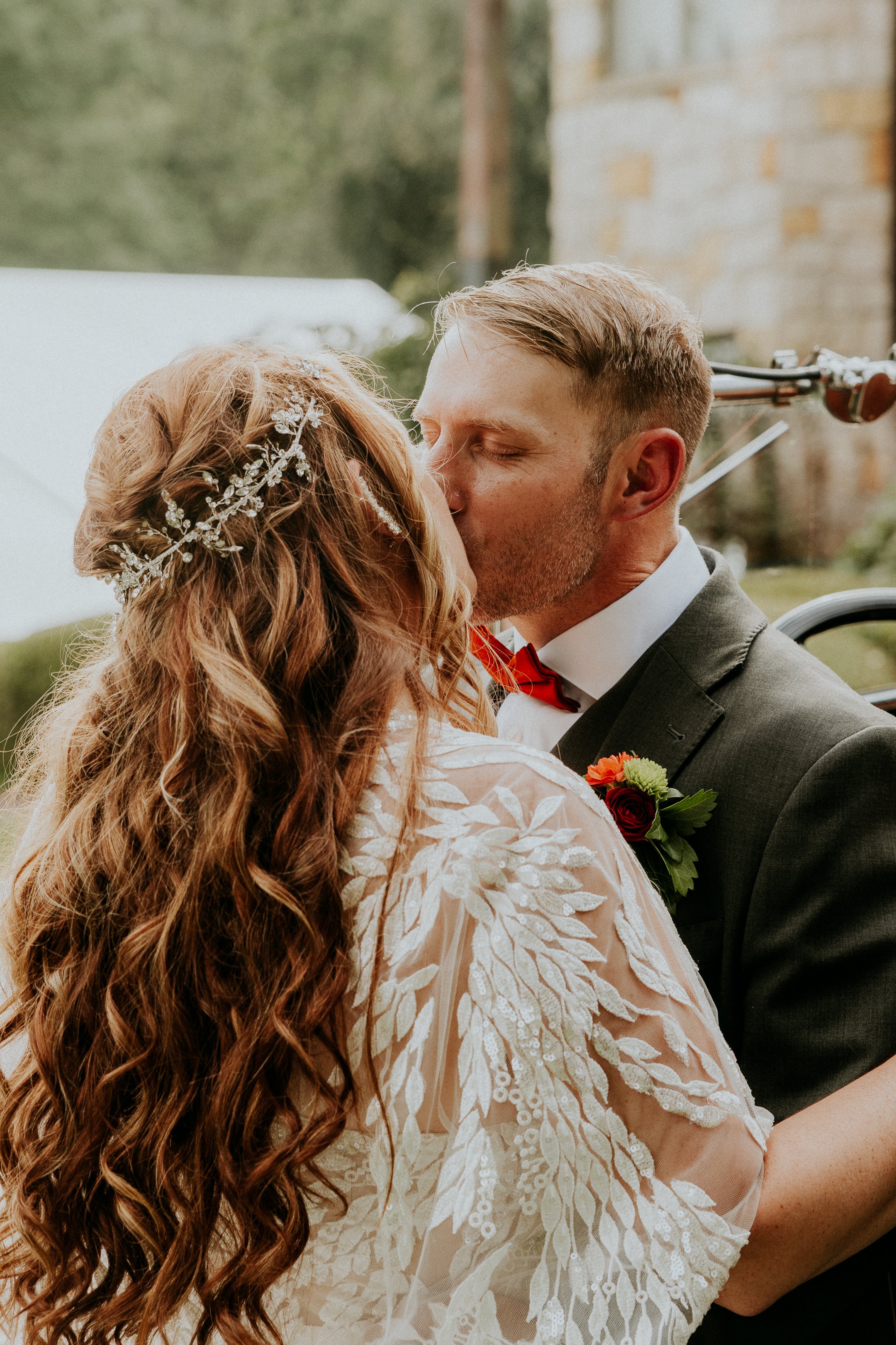 A couple sharing a kiss at their wedding outdoors with a wedding dress and tuxedo. The bride has long, curly hair with a decorative hairpiece. The groom has short, styled hair and a boutonniere with red and green flowers.