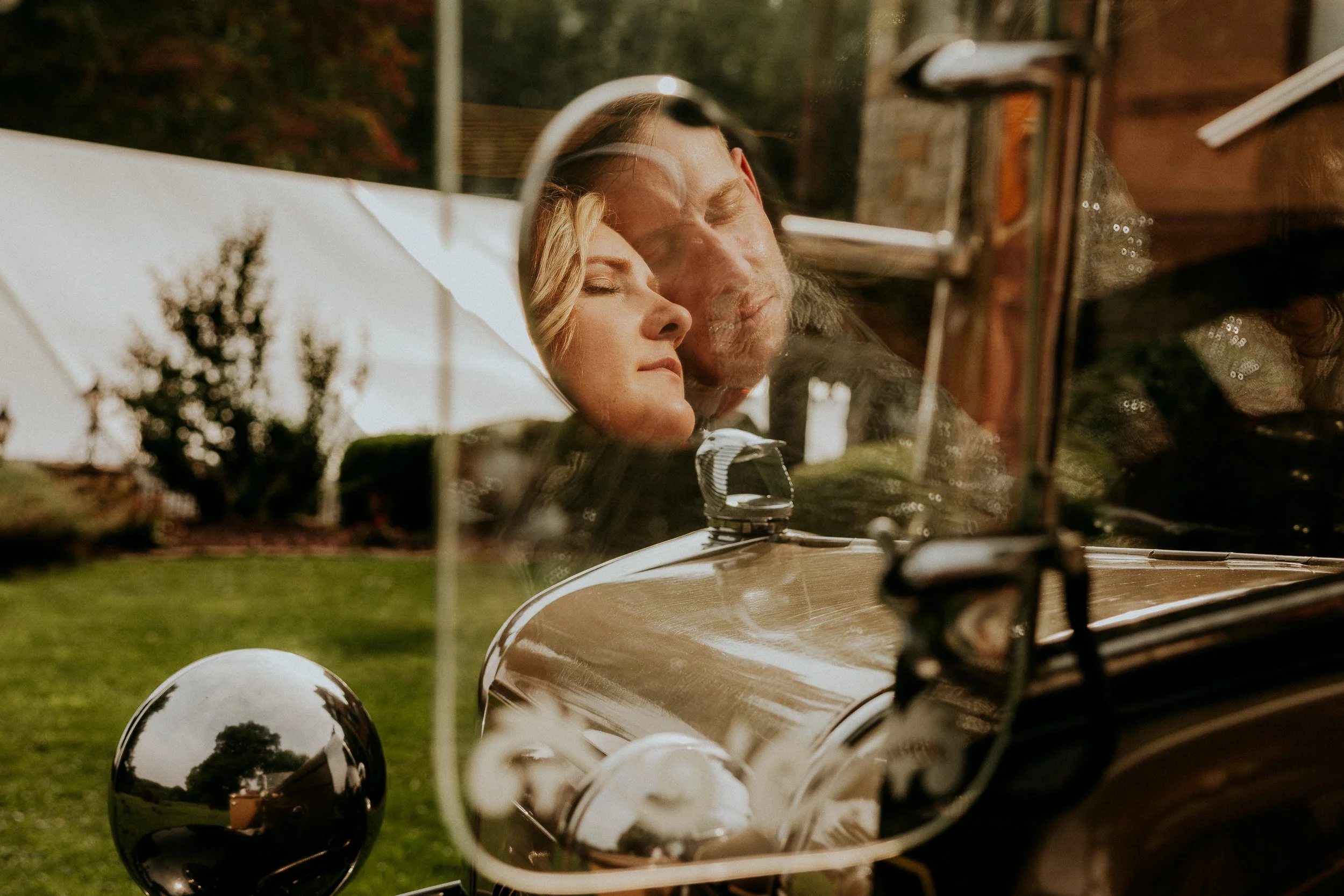 Close-up of a vintage car's side mirror reflecting a woman and man with closed eyes, surrounded by trees and greenery, with additional reflections of trees in the car's shiny surface.