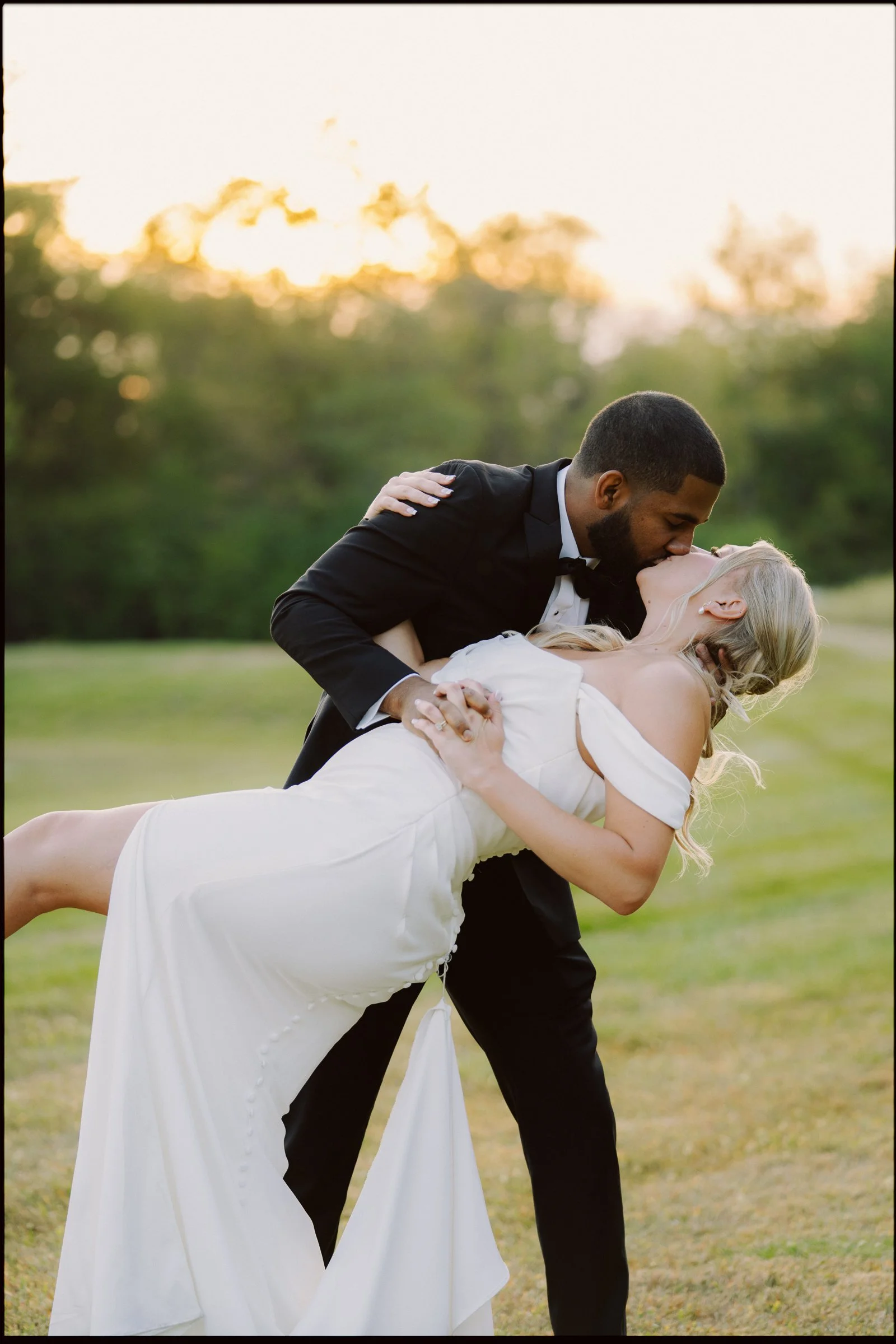 A man in a tuxedo is dipping a woman in a white wedding dress in an outdoor setting during sunset, sharing a kiss.