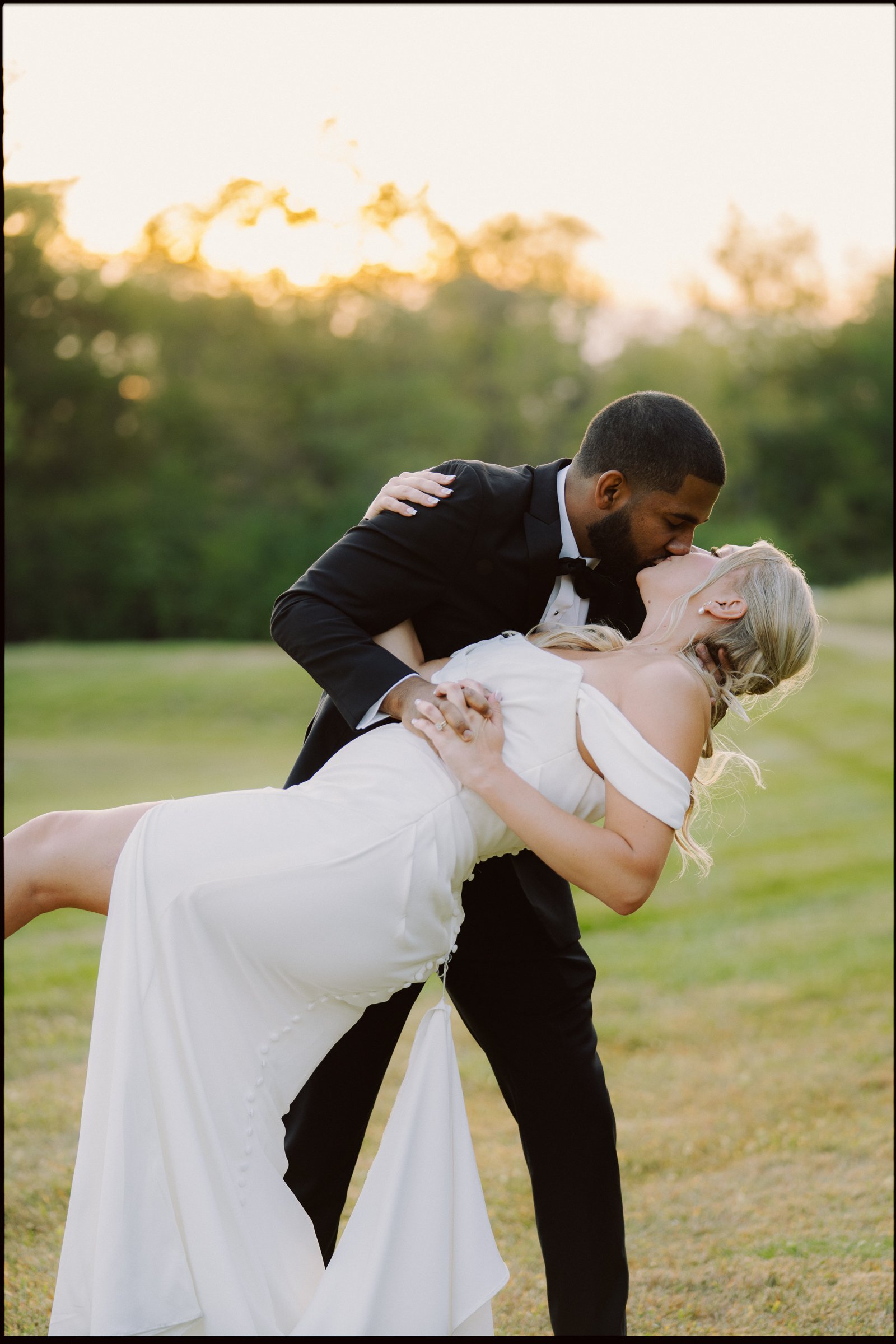 A wedding couple outdoor during sunset. The groom, dressed in a black tuxedo, dips the bride, who is in a white wedding dress, and kisses her. The background is a blurred natural setting with trees and sunlight.
