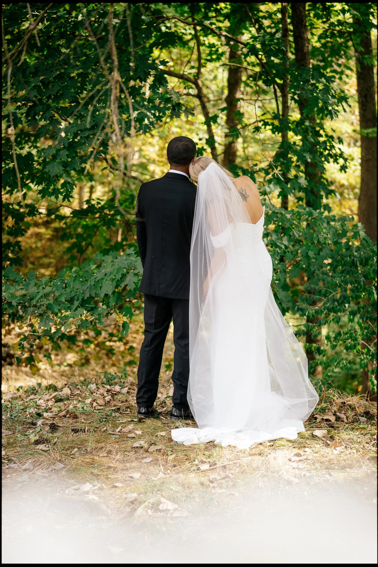 A newlywed couple standing close together in a wooded outdoor setting, with their backs to the camera. The groom is in a black suit, and the bride is in a white wedding gown with a veil, leaning her head on the groom's shoulder.