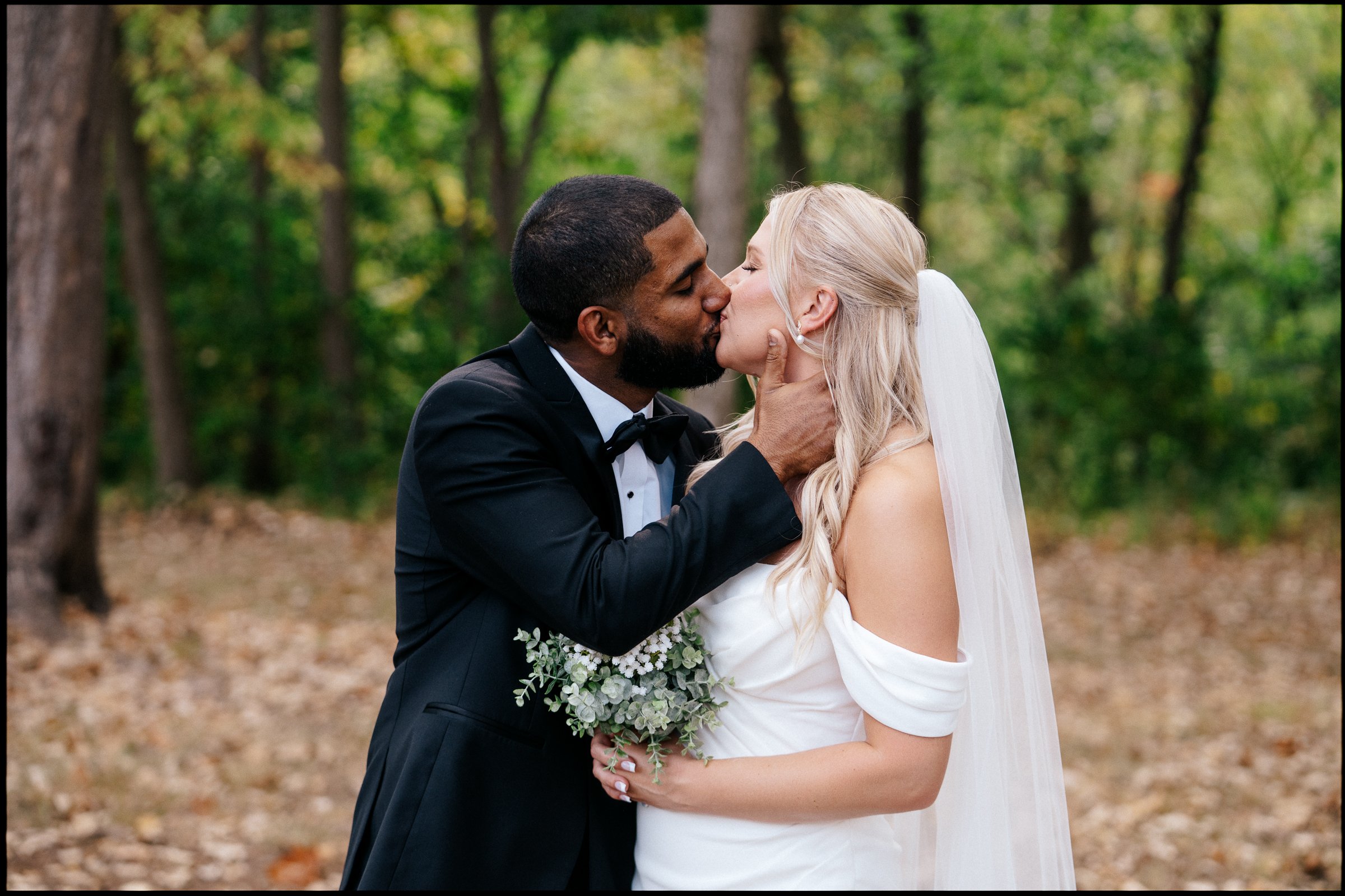 A bride and groom share a kiss outdoors in a wooded area. The groom wears a black tuxedo and the bride wears a white wedding dress with a veil. She holds a bouquet of white flowers and greenery. Trees with green leaves surround them.