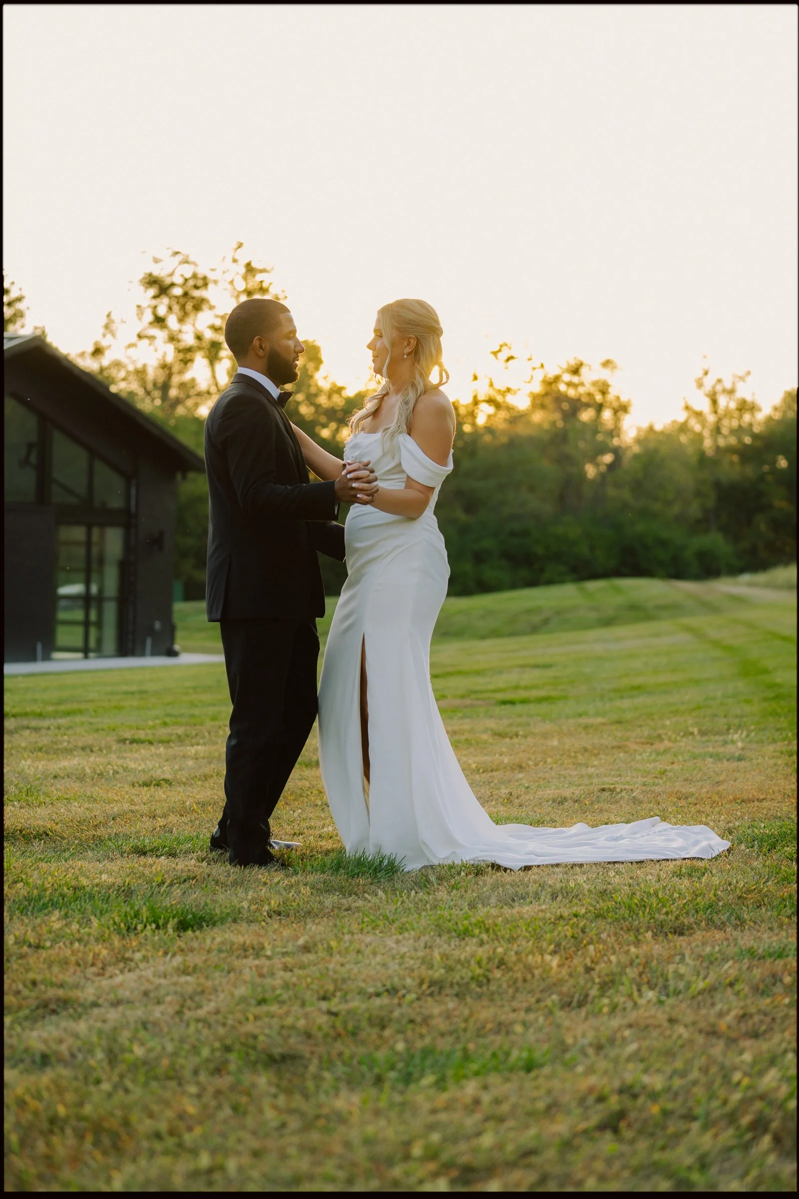 A bride and groom standing on a grassy field during sunset, sharing a dance.