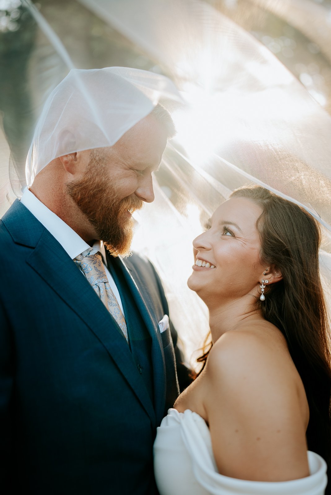 A bride and groom looking at each other under a clear umbrella, sharing a joyful moment during their wedding.
