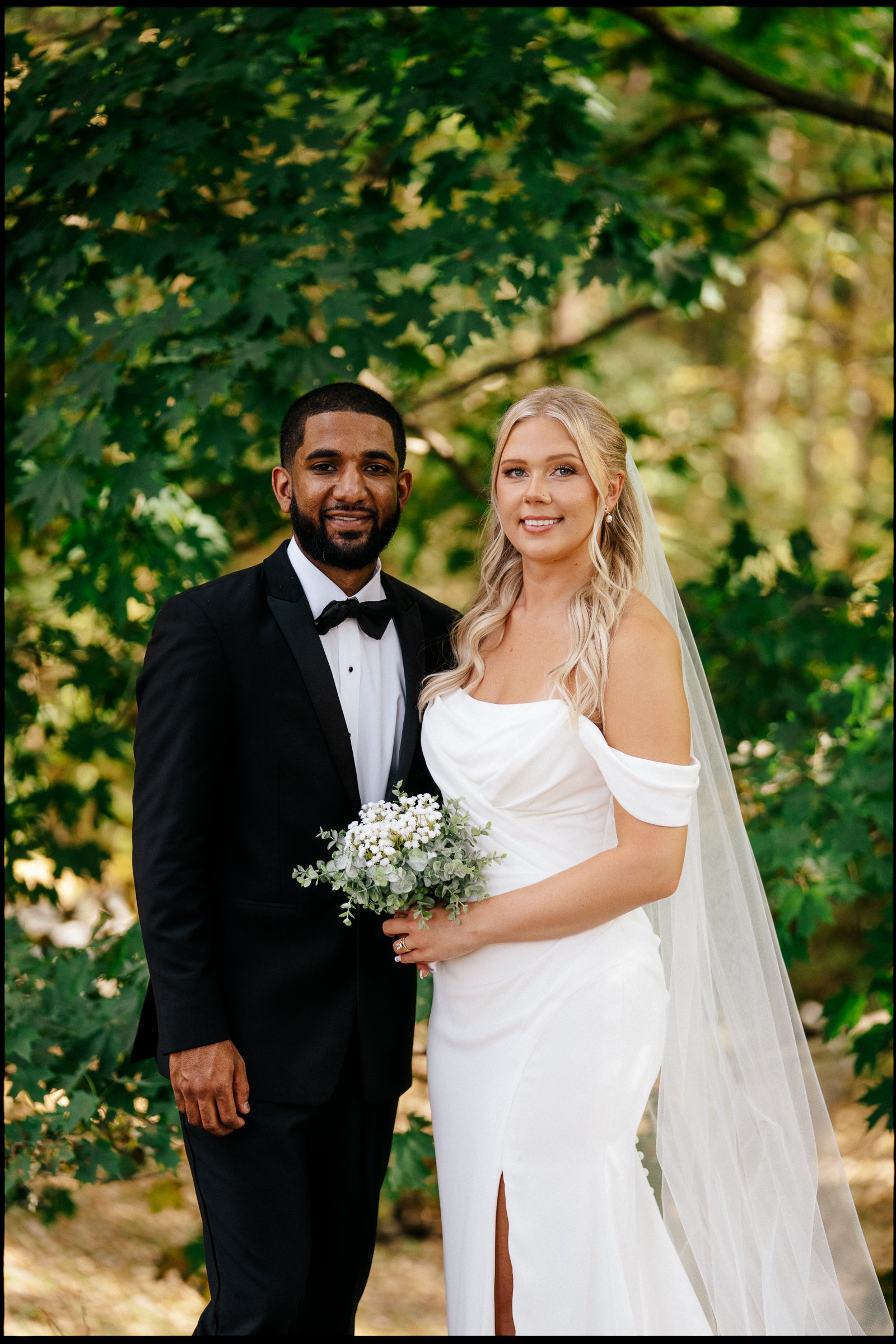 A newlywed couple poses outdoors in front of green leaves. The groom is wearing a black tuxedo with a bow tie, and the bride is in a white wedding dress holding a bouquet of white flowers. The bride has long blonde hair and the groom has short dark h