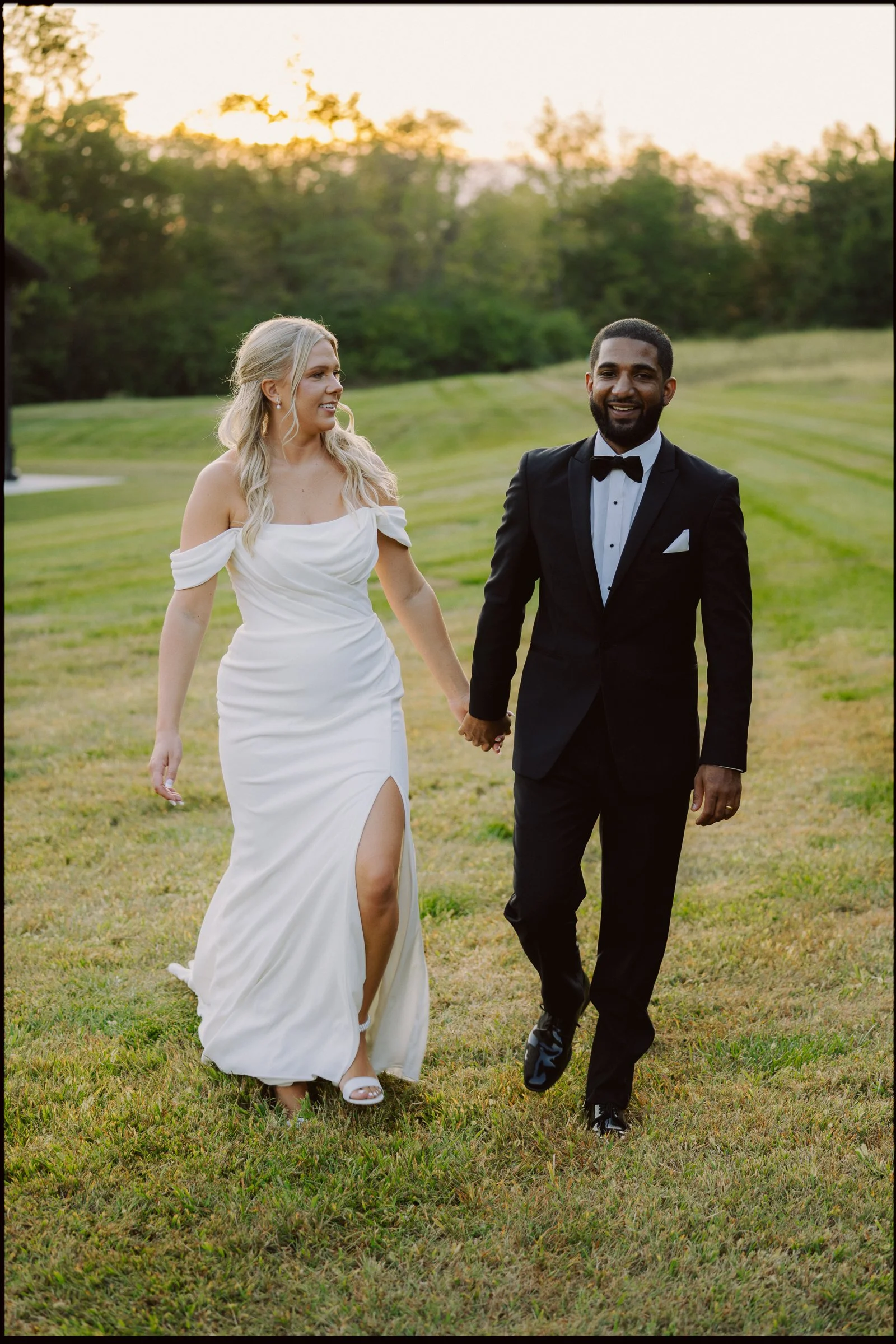 A newlywed couple holding hands and walking on a grassy field during sunset, with trees in the background. The woman is wearing a white wedding dress with off-the-shoulder sleeves and a slit, and the man is dressed in a black tuxedo with a bow tie.