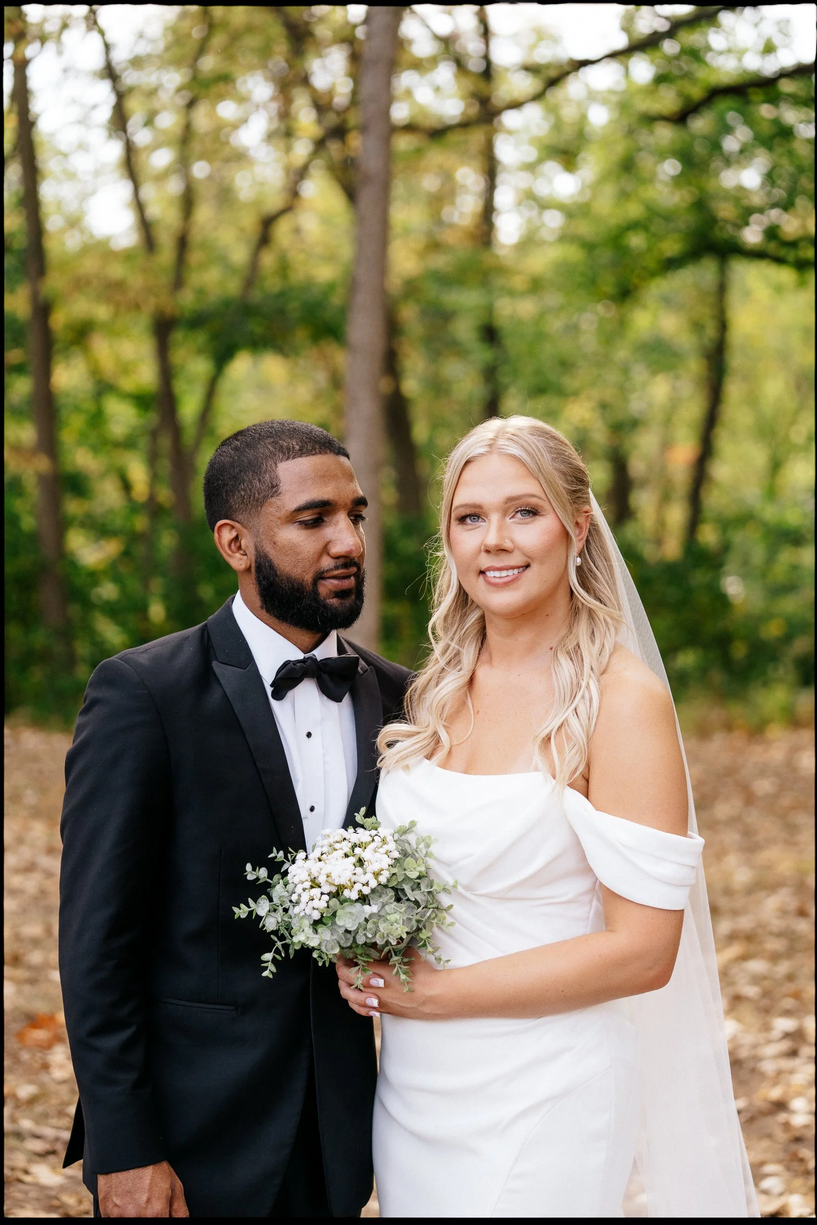A bride and groom standing outdoors in a forested area for a wedding, with green trees in the background. The groom is wearing a black tuxedo with a bow tie, and the bride is wearing a white wedding dress, holding a bouquet of flowers and smiling.