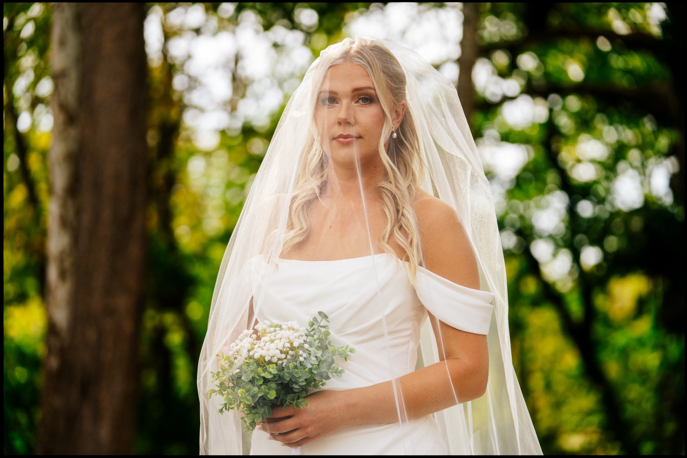 A bride with long blonde hair in a loose updo, wearing a white off-the-shoulder wedding dress and veil, holding a bouquet of white flowers and greenery, standing outdoors in a wooded area with dappled sunlight.