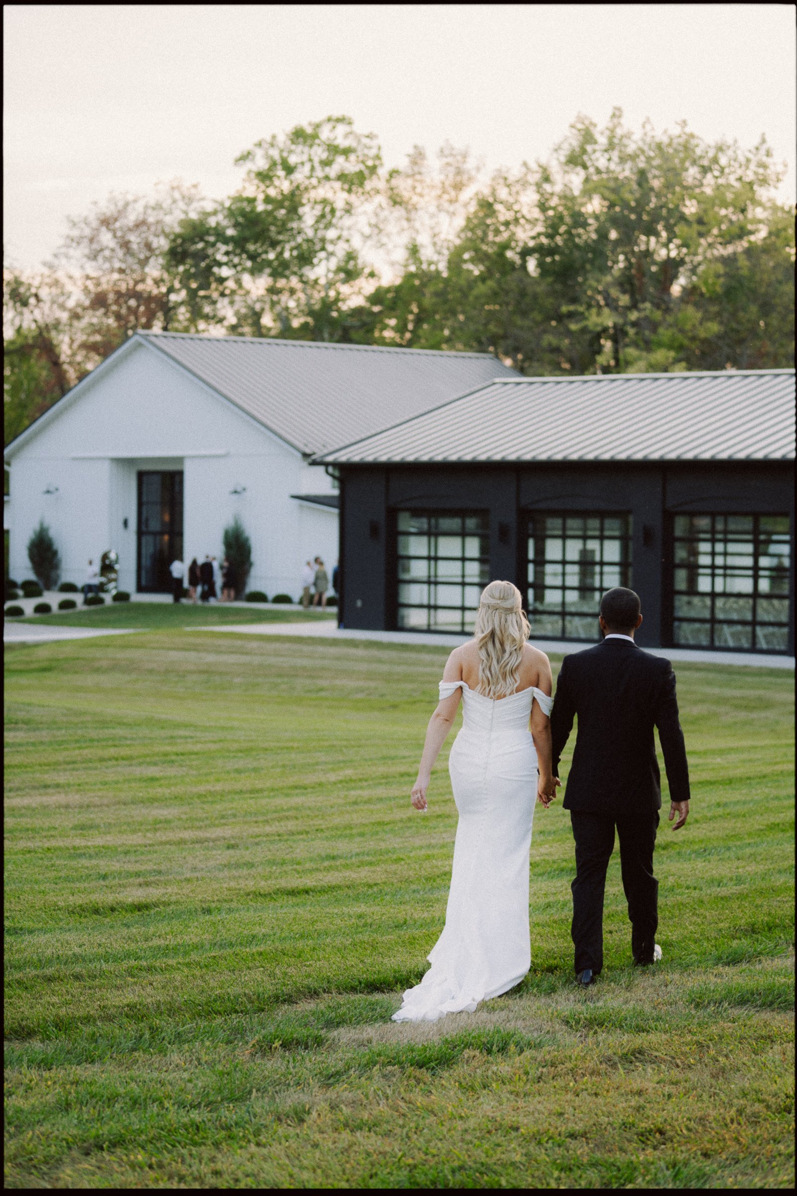 A bride in a white wedding dress and a groom in a black suit walking hand-in-hand across a well-manicured lawn toward a modern white and black building, with trees in the background and people gathering outside.