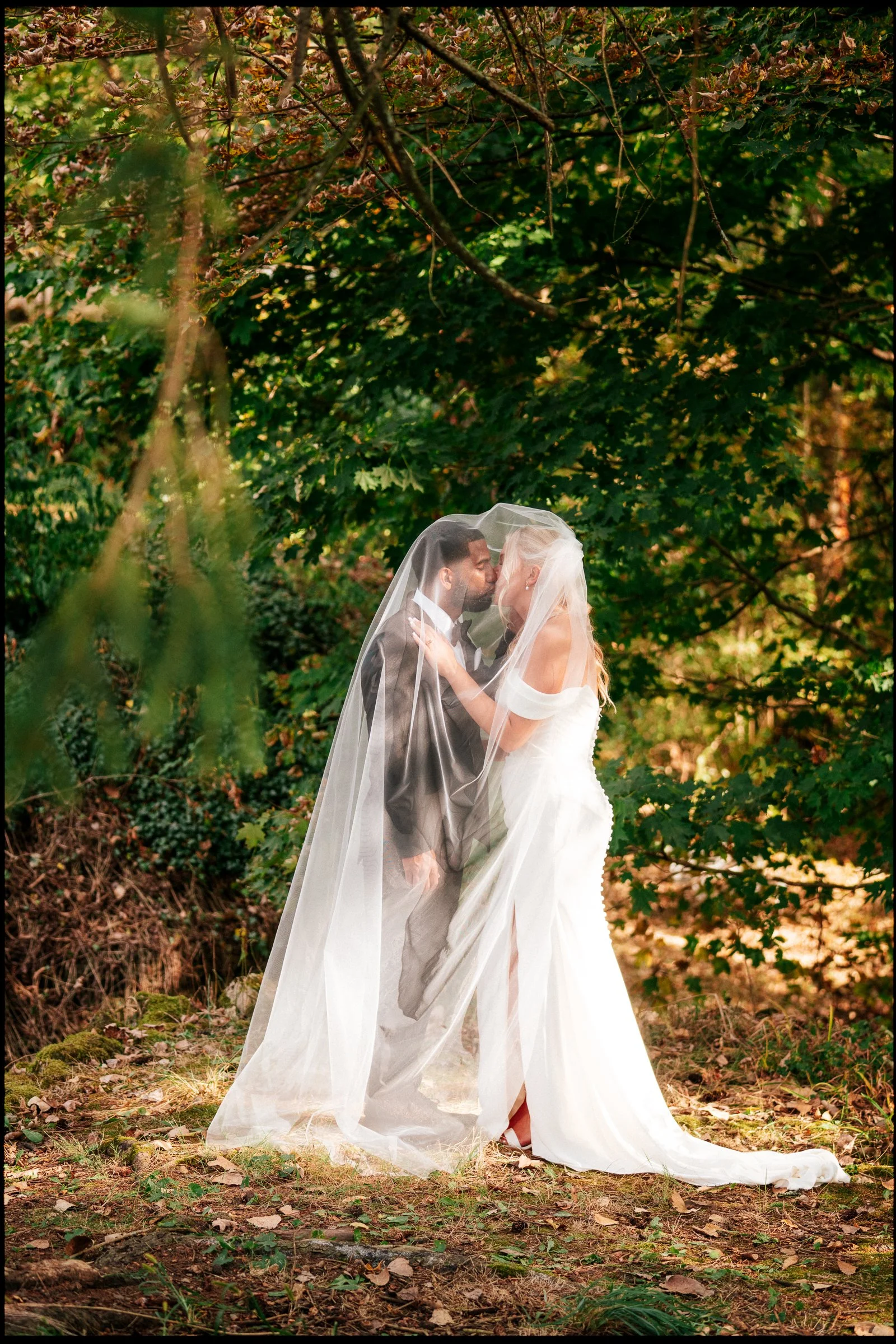 A bride and groom are standing close together, performing a kiss in a forested area with lush green trees and foliage around them. The bride wears a white wedding gown and veil, while the groom wears a dark suit.