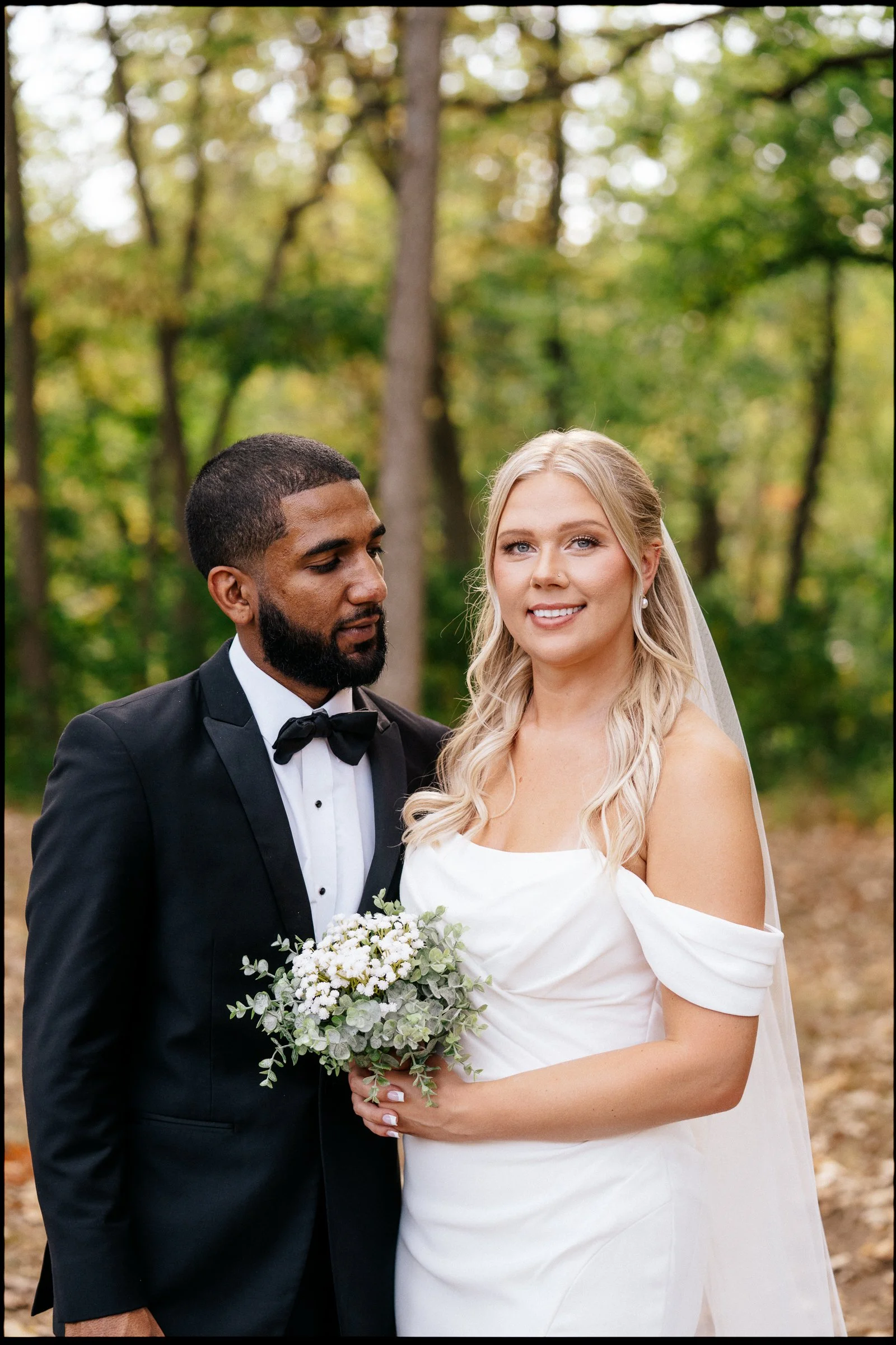 A newlywed couple standing outdoors in a forested area with blurred trees in the background. The groom, wearing a black tuxedo with a bow tie, is looking at the bride. The bride, with long blonde hair in loose waves, is smiling at the camera and hold