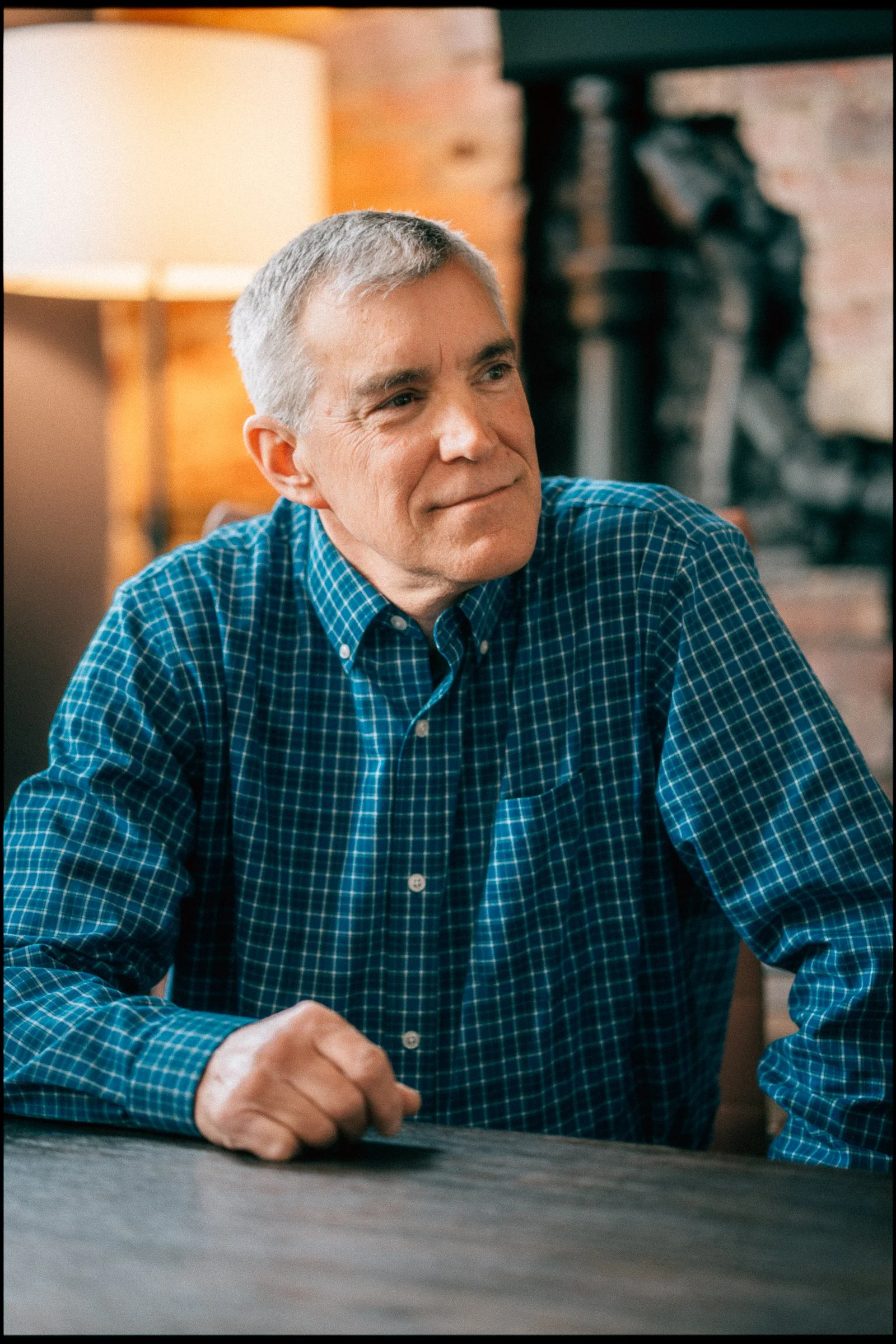 A middle-aged man with short gray hair wearing a blue checkered shirt, sitting at a table with a contemplative expression, in a cozy indoor setting with a lamp and brick wall in the background.