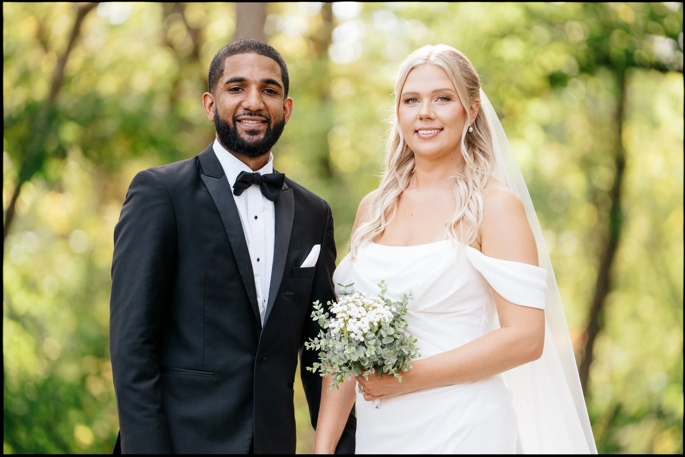 A bride and groom stand together outdoors, smiling, with the bride holding a bouquet of white flowers and greenery. The groom is wearing a black tuxedo with a black bow tie, and the bride is in a white wedding dress with off-the-shoulder sleeves and 