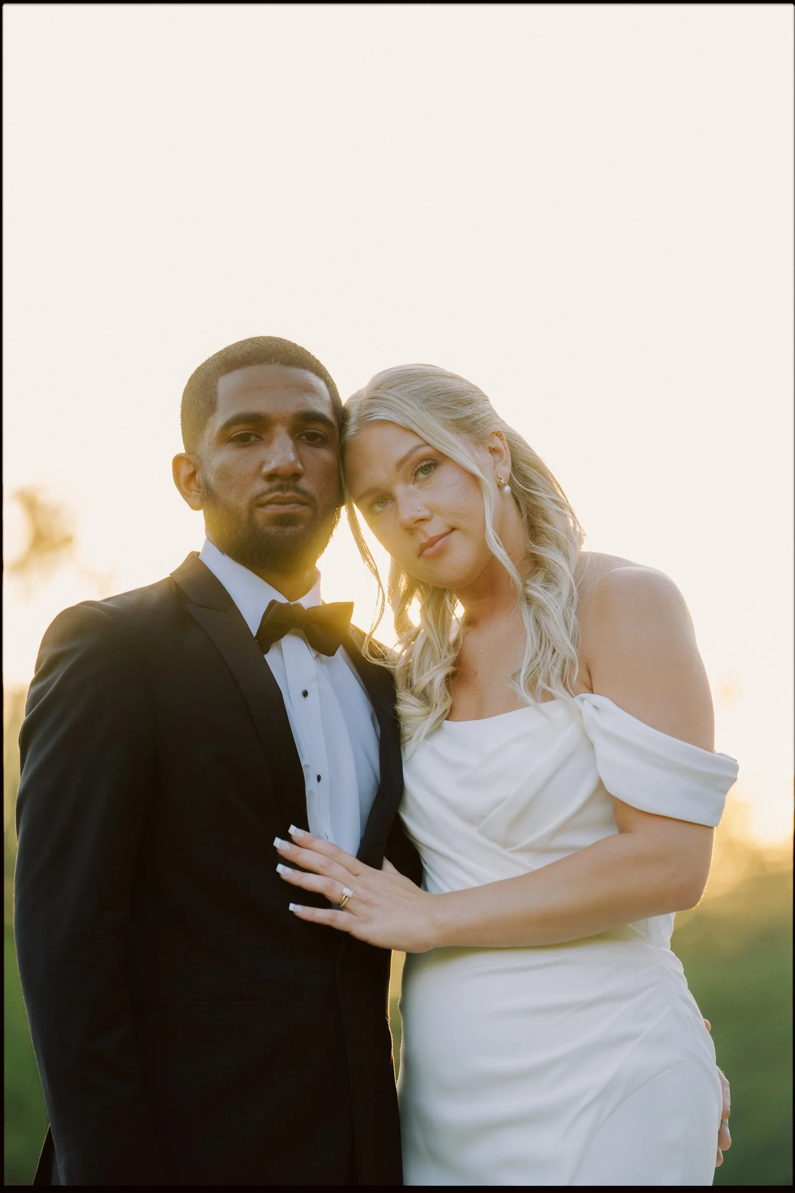 A couple at their wedding, outdoors during sunset, with the groom in a black tuxedo and the bride in a white off-shoulder wedding dress. They are close together, with the bride's hand on the groom's chest, showing wedding rings.