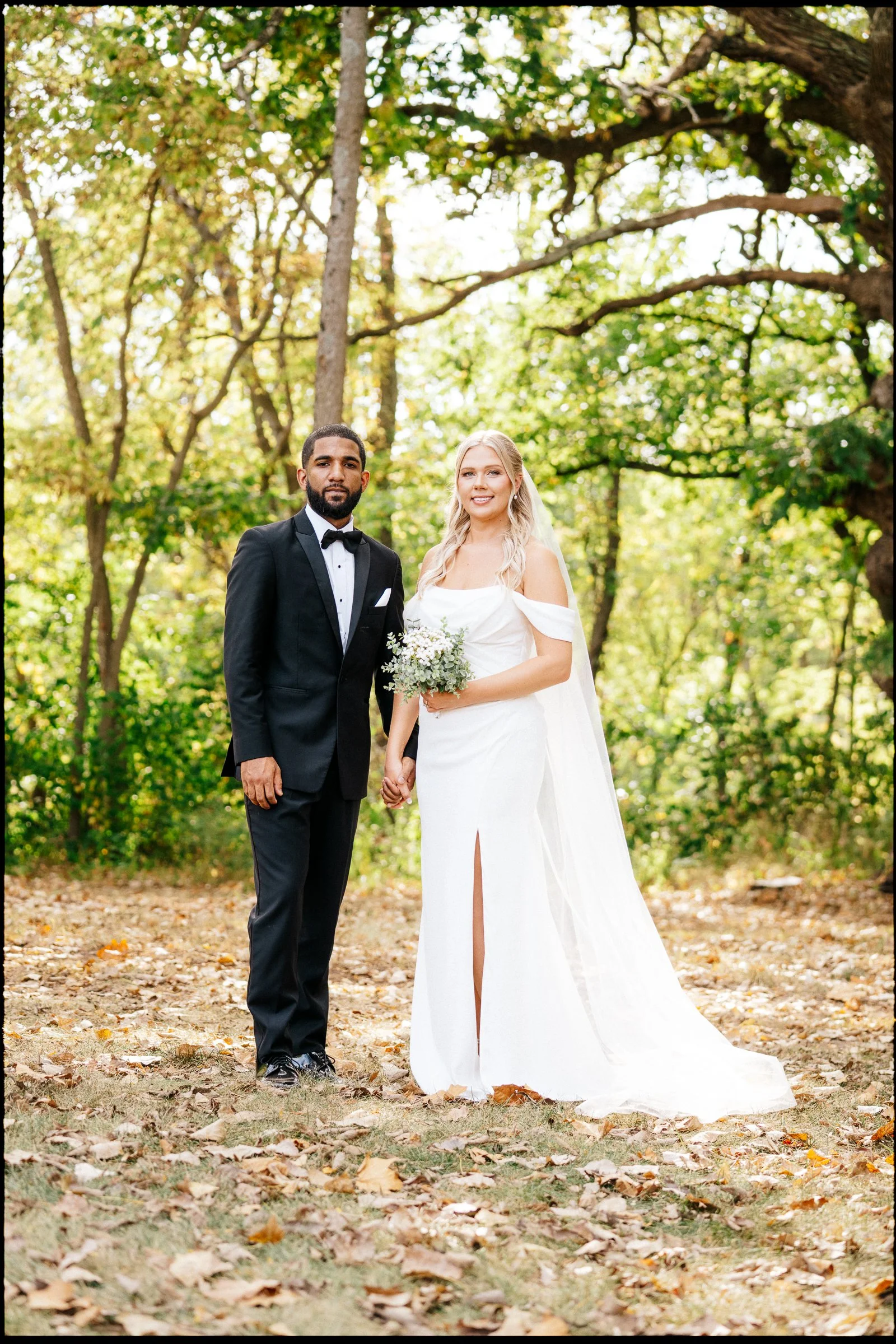 A bride and groom standing outdoors in a wooded area with fallen leaves on the ground, holding hands and looking at the camera. The groom is dressed in a black tuxedo with a bow tie, and the bride is wearing a white wedding dress holding a bouquet of
