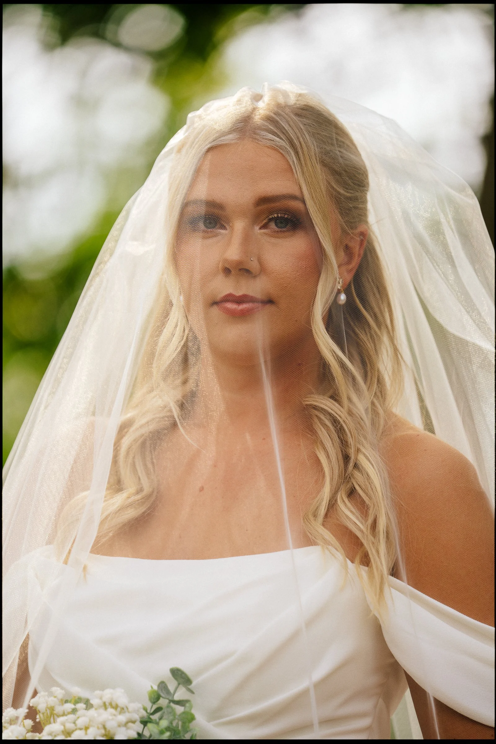 A bride with long blonde hair and light makeup, wearing a strapless white wedding dress and pearl earrings, standing outdoors with a sheer veil over her head, holding a bouquet of small white flowers and greenery.
