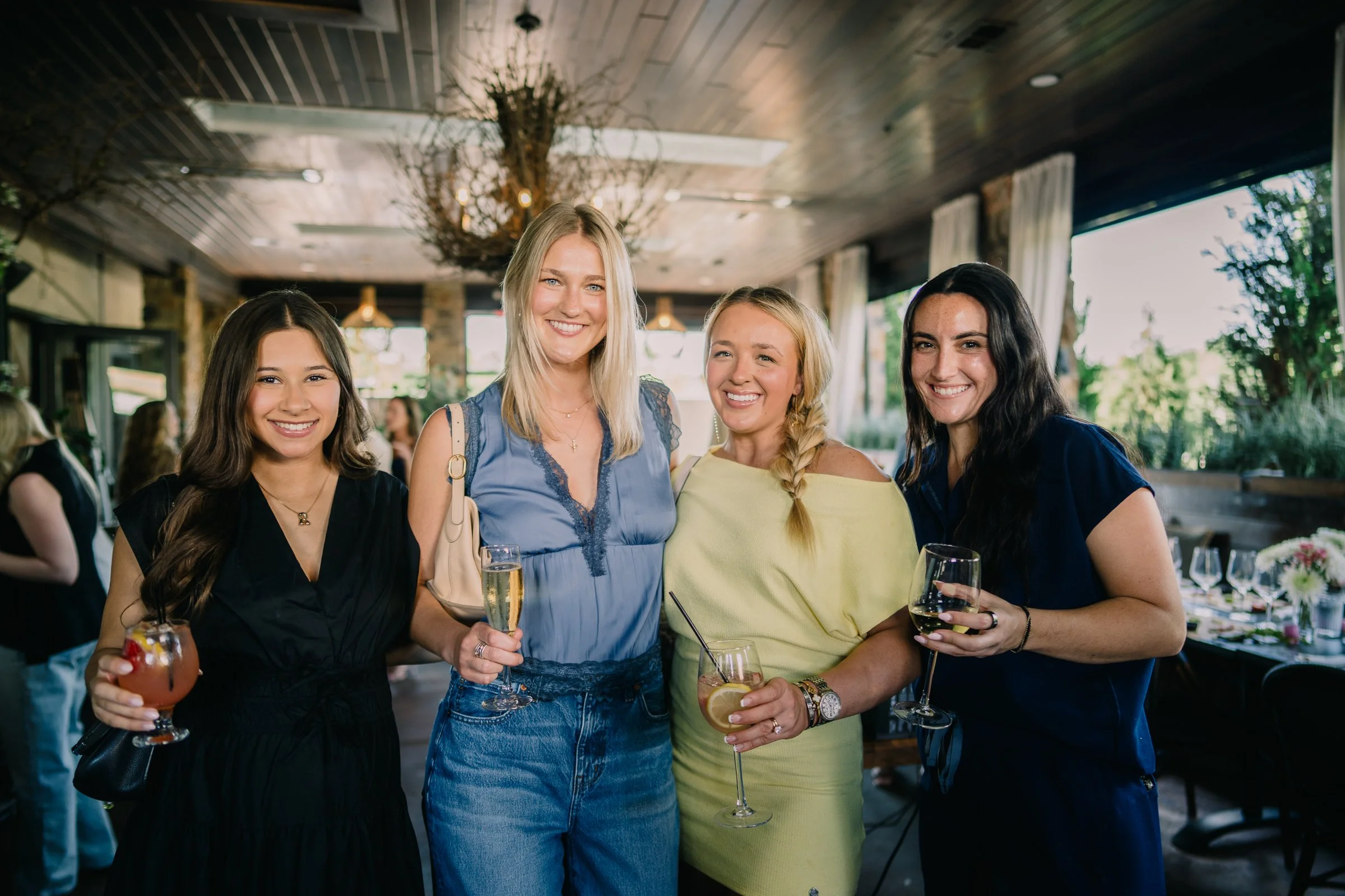 Four women smiling and holding drinks at a social gathering in a warmly lit indoor venue with large windows and greenery outside.