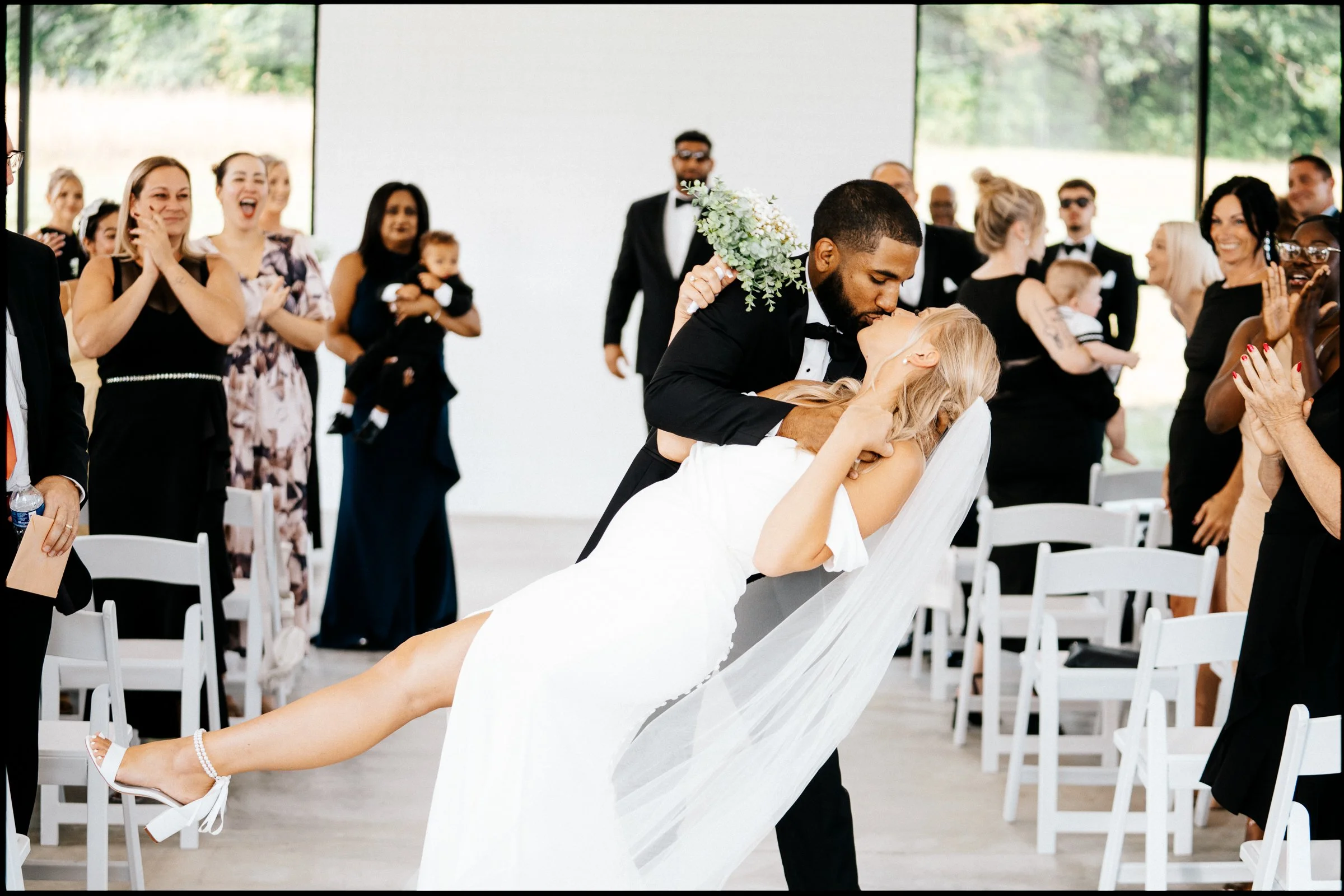 A groom is kissing a bride who is leaning back, while guests in formal attire watch and smile at an indoor wedding ceremony.