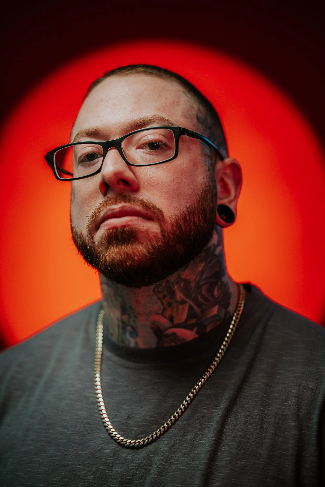 Close-up of a man with glasses, tattoos on neck, beard, wearing a chain, against a red background.
