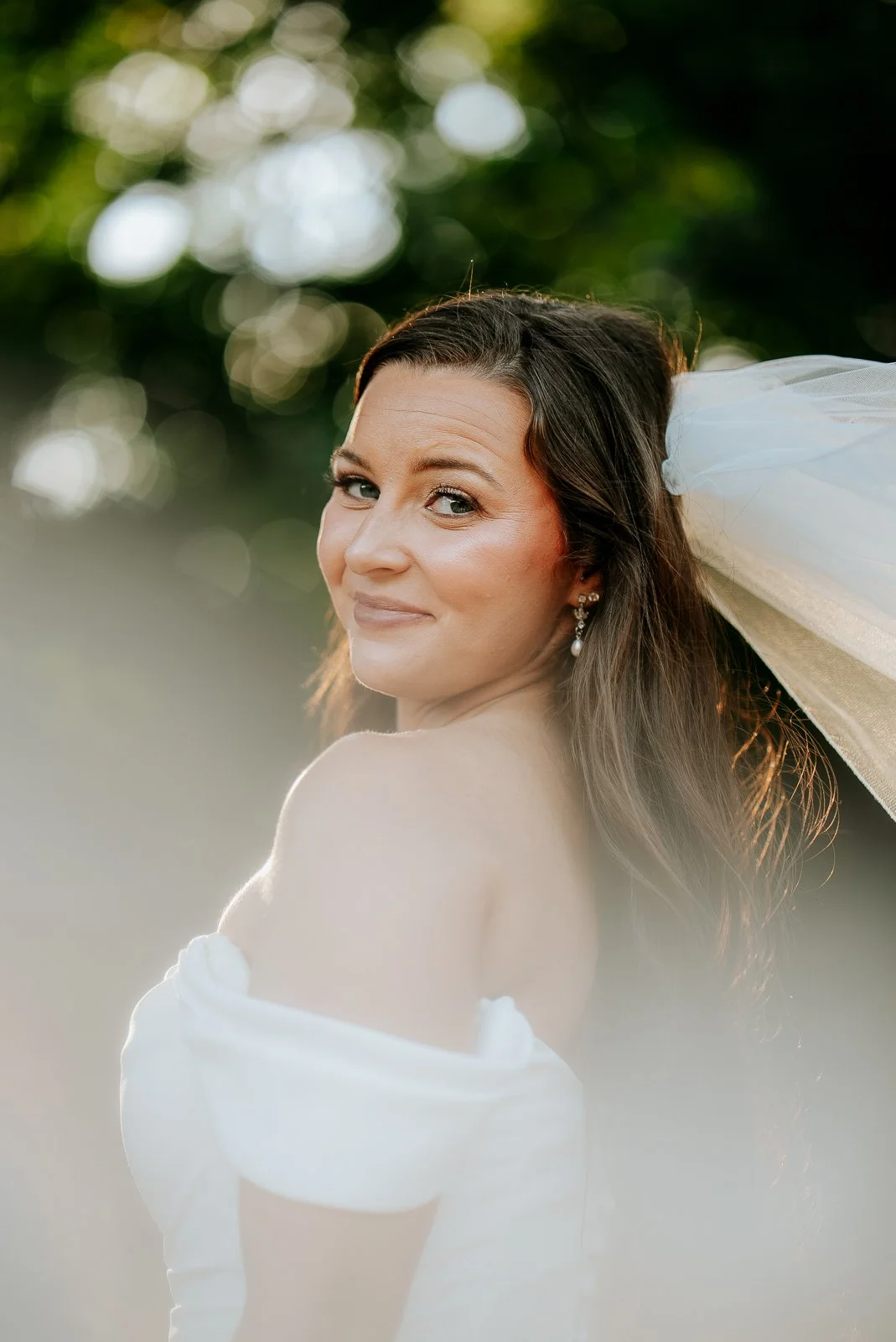 A woman with long brown hair, earrings, and a white off-shoulder dress, looking over her shoulder at the camera, outdoors with blurred greenery background.