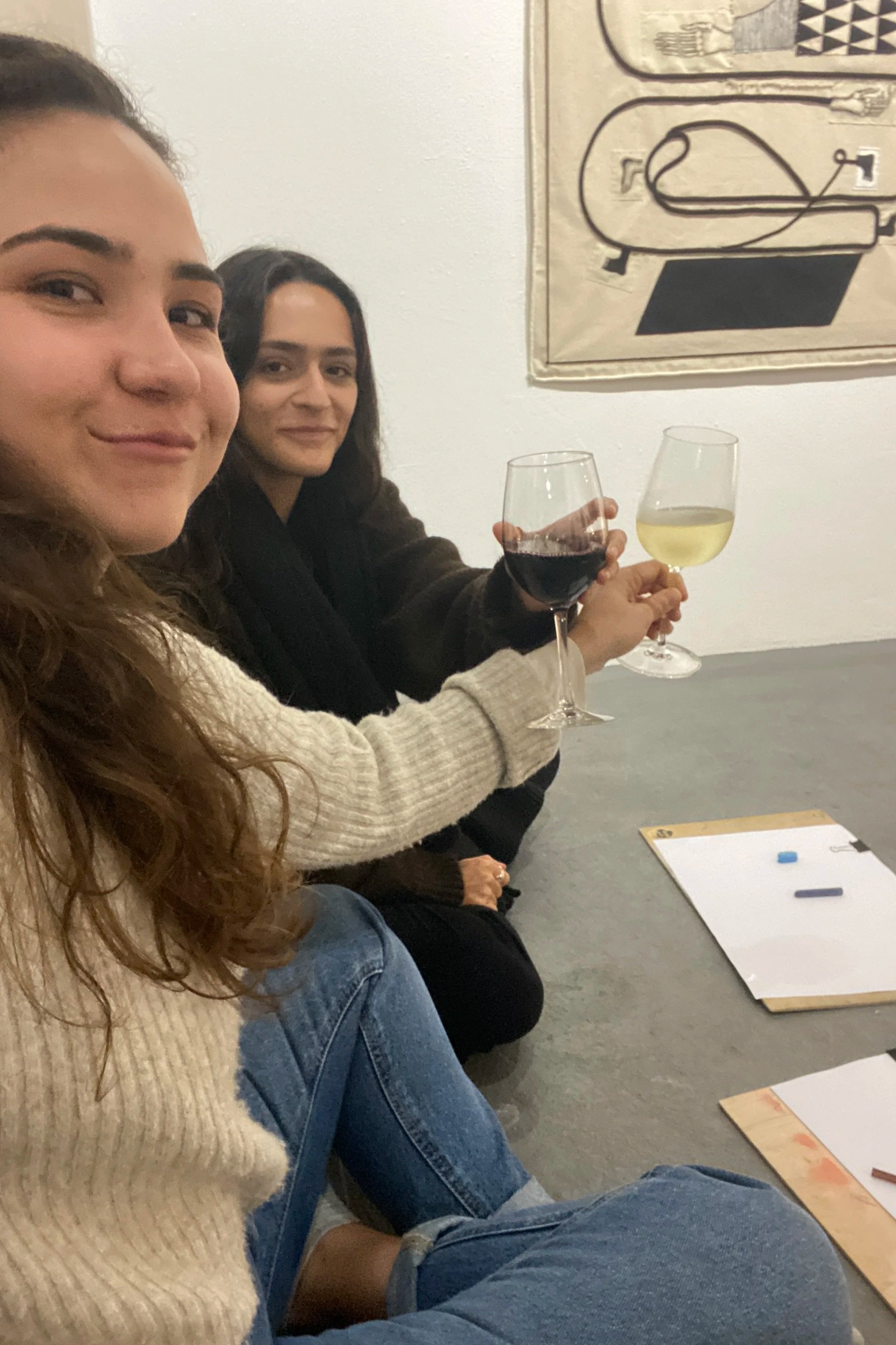 Two women sitting on the floor at a table, holding glasses of wine, with art and papers on the wall and table.