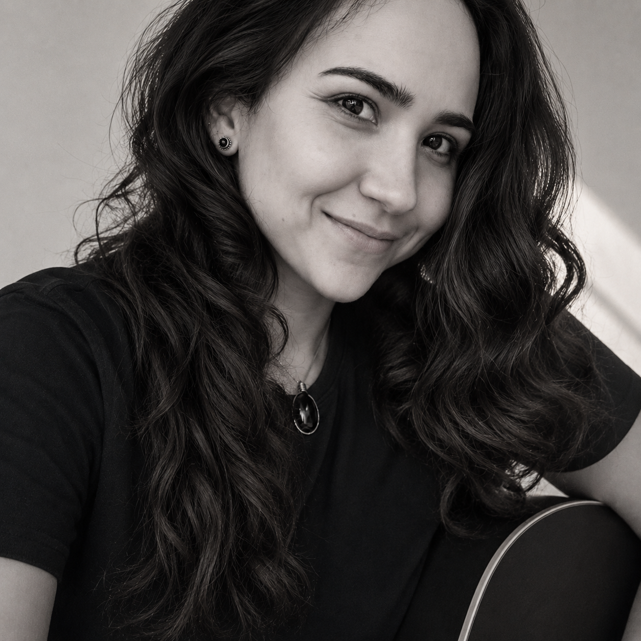 Black-and-white photo of a young woman with wavy dark hair, smiling gently, wearing a necklace, earrings, and a dark shirt, with part of a guitar visible.