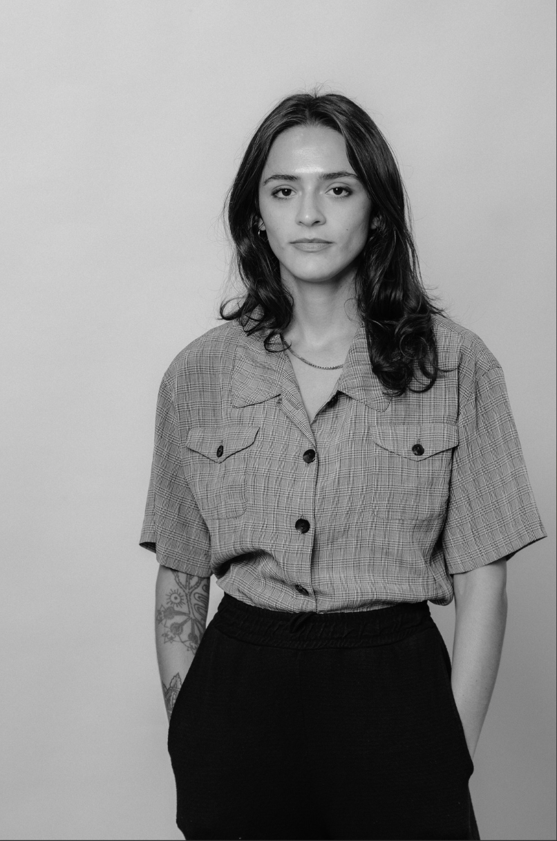 A young woman with shoulder-length wavy hair standing against a plain background, wearing a checkered short-sleeve shirt, dark pants, and a necklace.