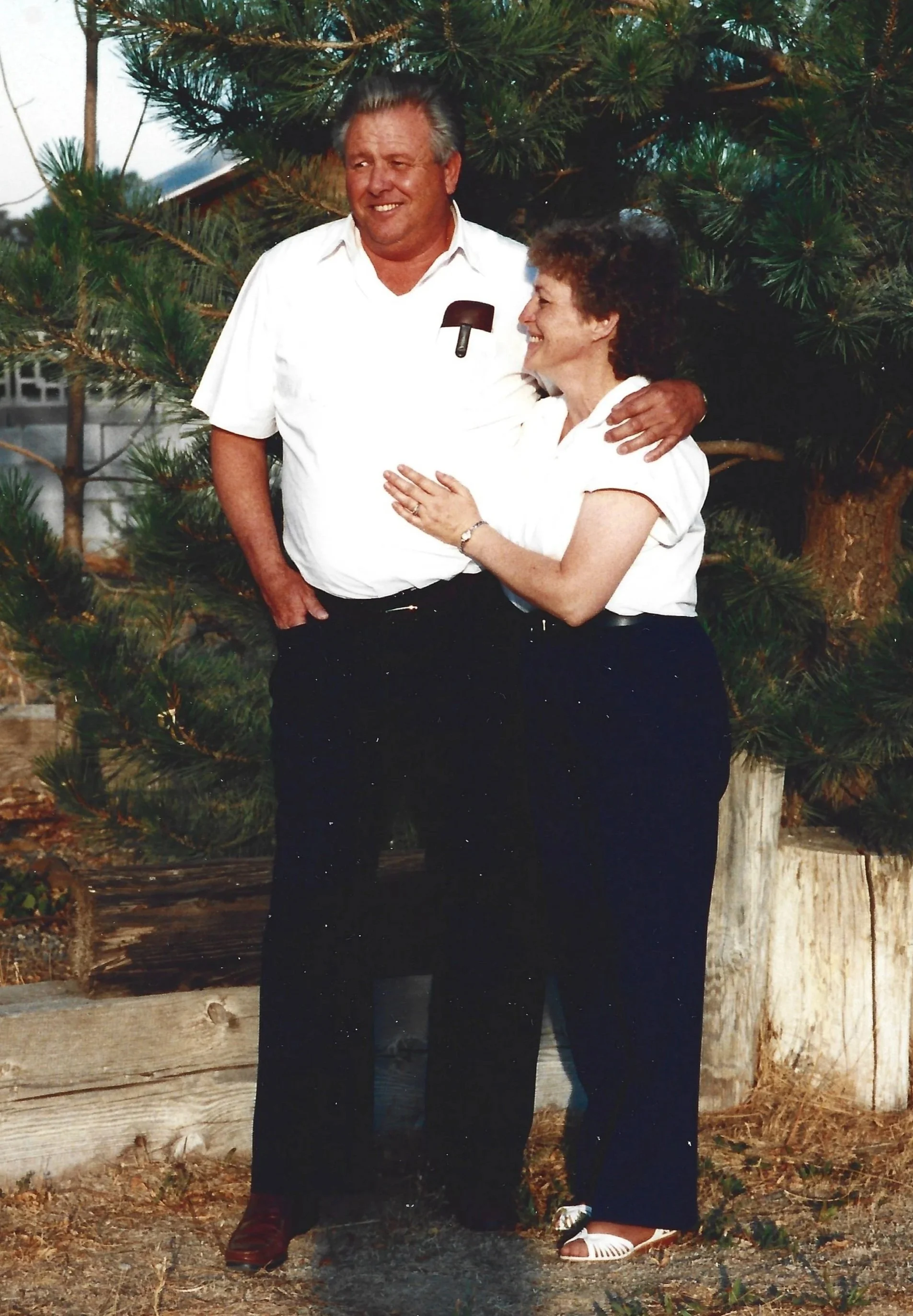 A man and woman are standing outdoors near a tree, smiling at each other. The man has gray hair and is wearing a white short-sleeve shirt and dark pants. The woman has curly hair and is wearing a white blouse and dark pants. They are embracing with the man's arm around the woman's shoulders. The background includes a green tree and a wooden fence.