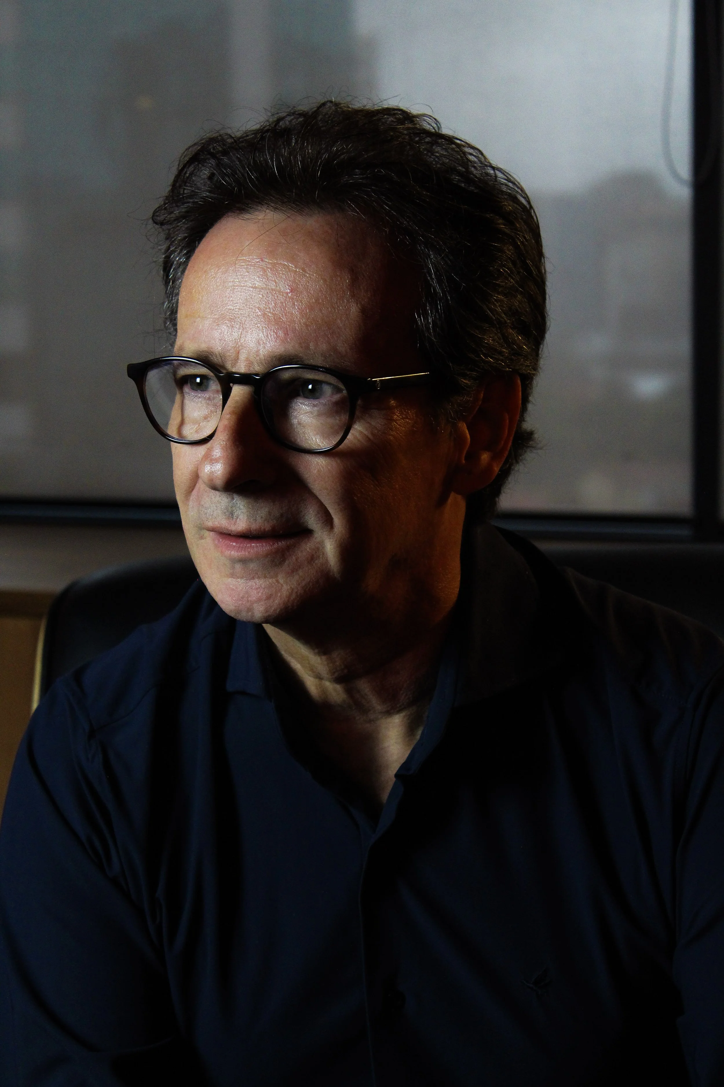 A man with dark hair and glasses looking to the side, wearing a dark shirt, in a dimly lit indoor setting.