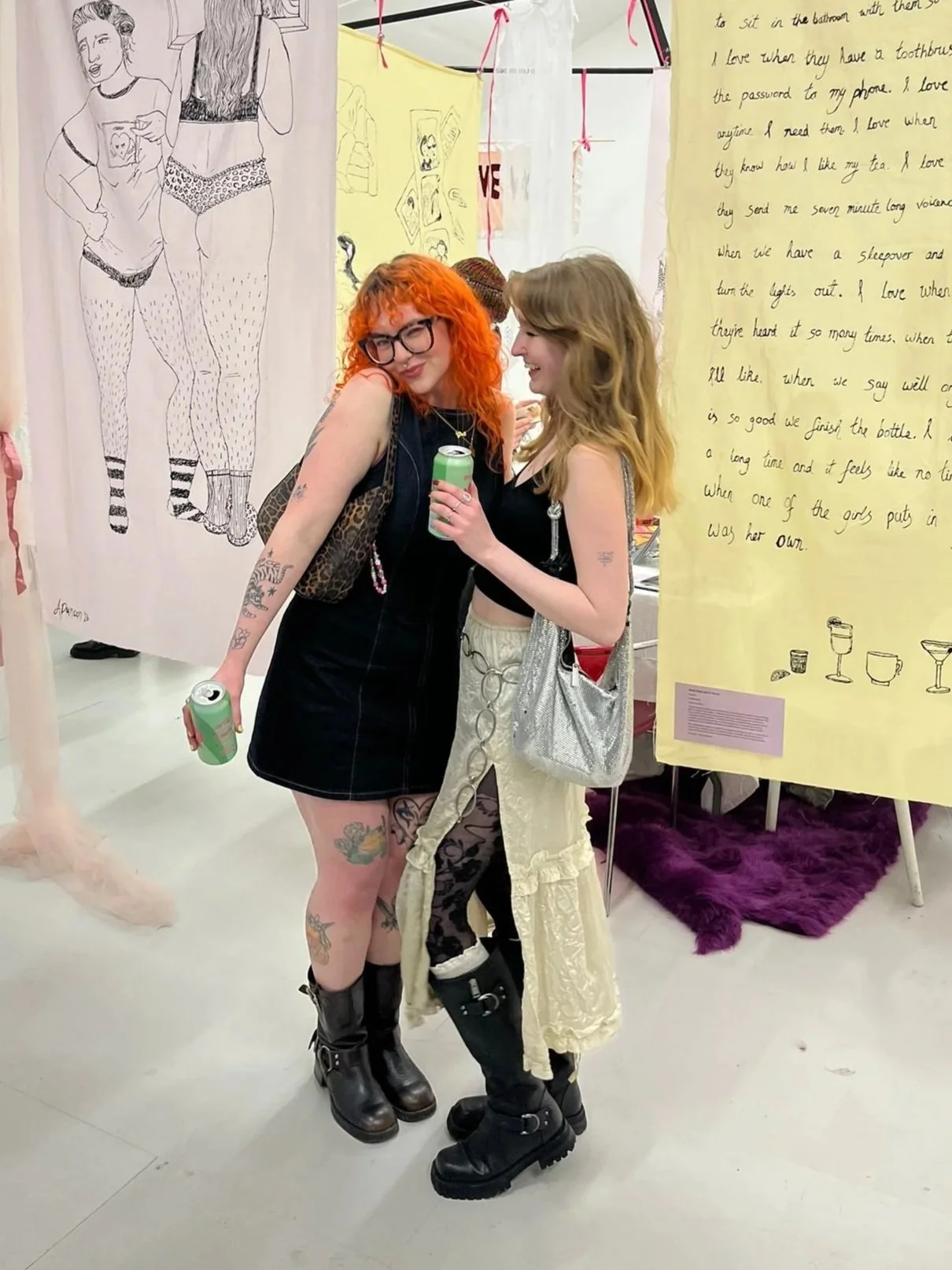 Two women with tattoos and colorful hair standing and posing indoors at an art exhibit or event, holding cans and smiling. Behind them are large illustrated panels and handwritten notes on display.