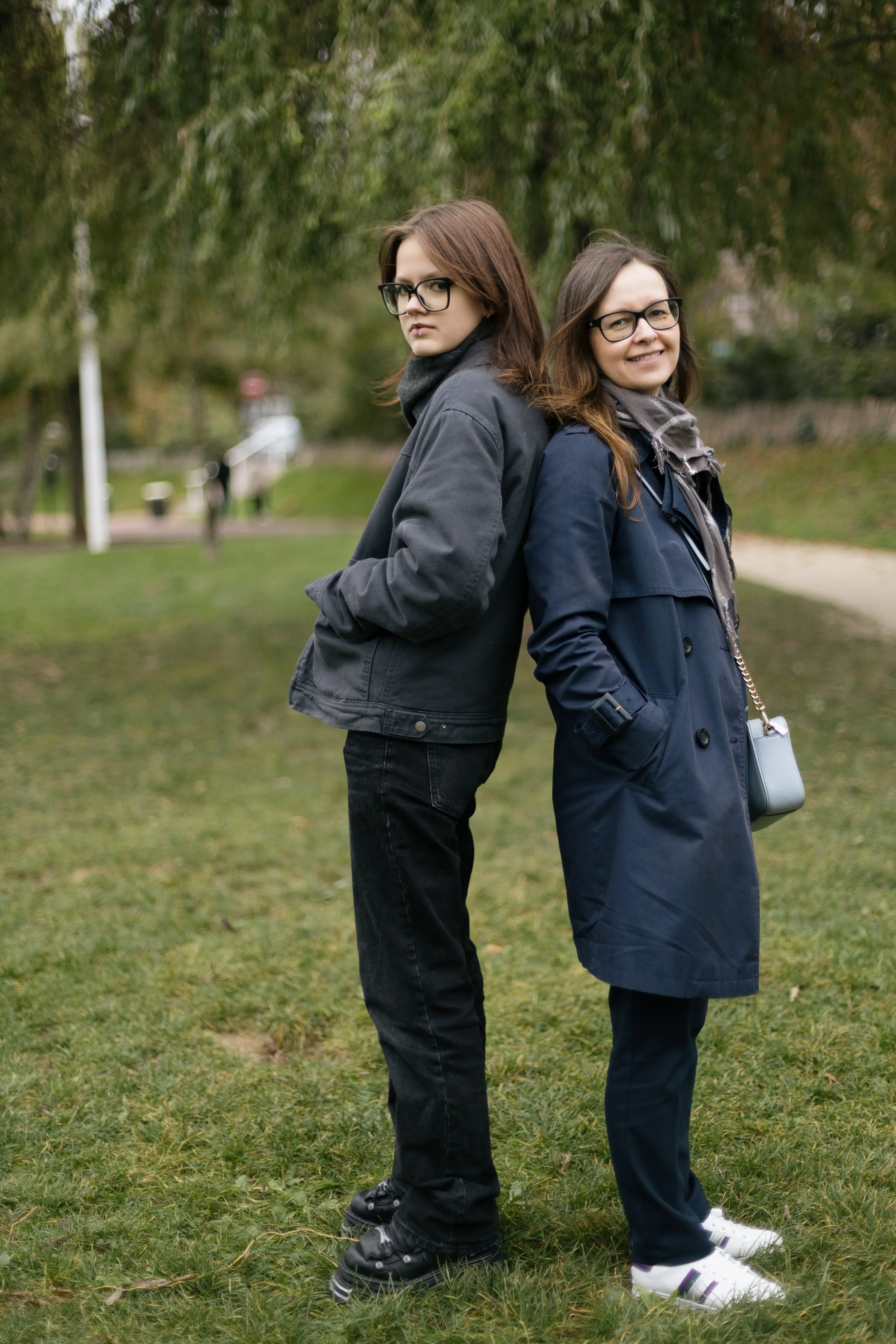 Two women standing back-to-back outdoors on a grassy area, both wearing glasses and dark-colored jackets, with one smiling and the other looking serious.
