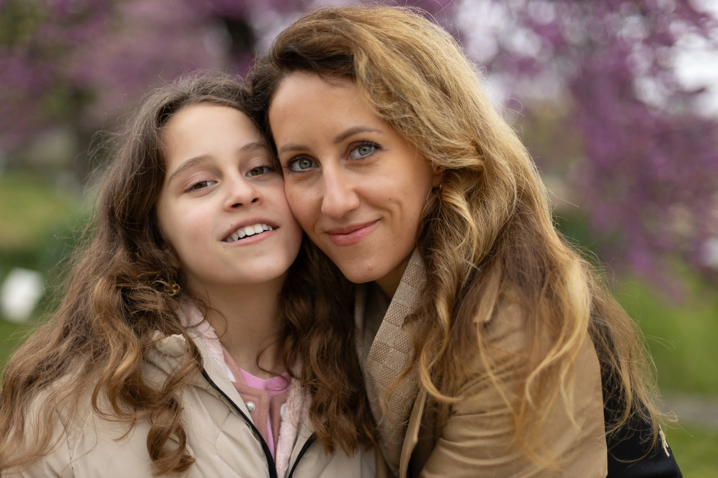 A woman and a girl with long, curly hair, smiling and close together outdoors with pink flowering trees in the background.