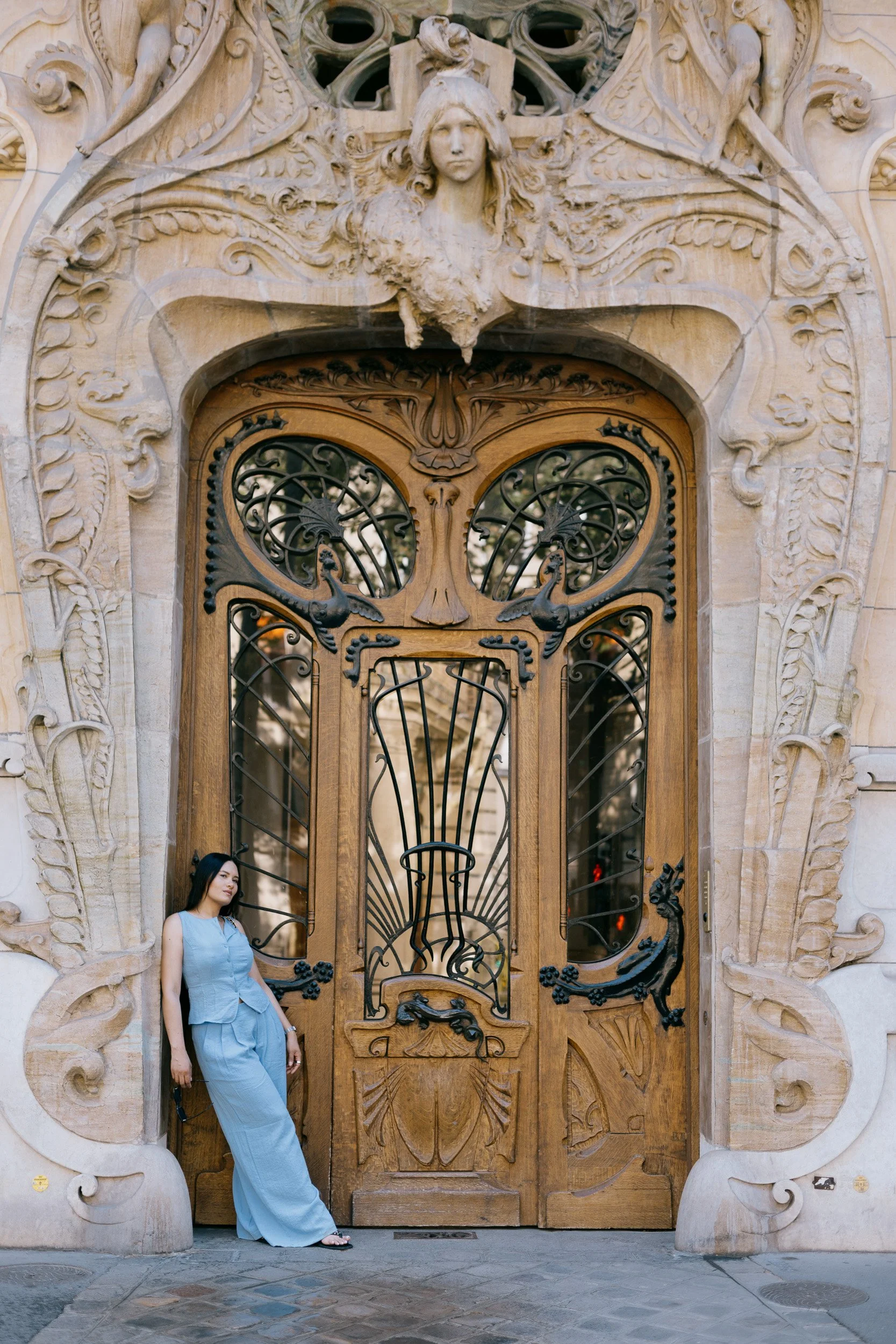 A woman wearing a light blue dress and black sandals is standing in front of an ornate wooden door with iron gate details and carved stone decorations, including a sculpted face at the top.