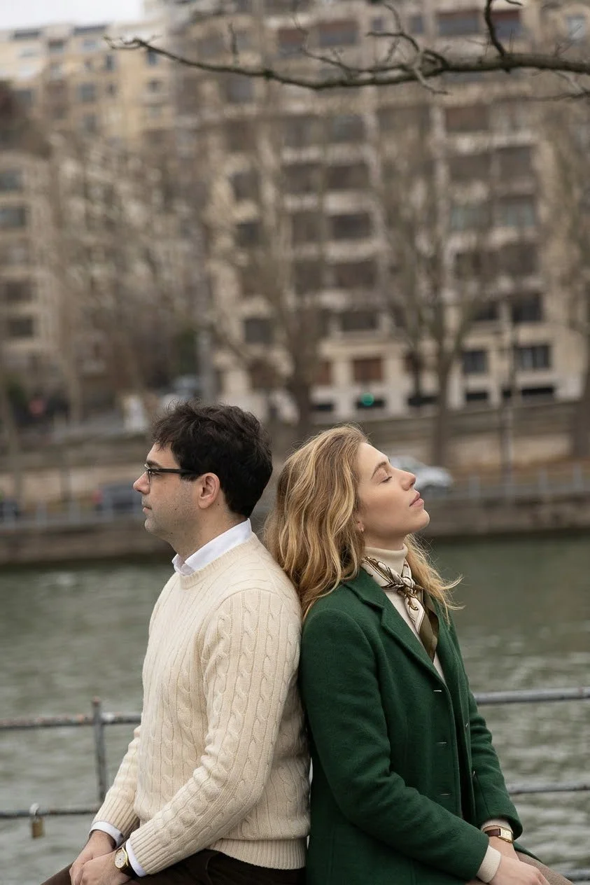 A man and a woman sit back-to-back near a river in an urban area, with trees and buildings in the background.