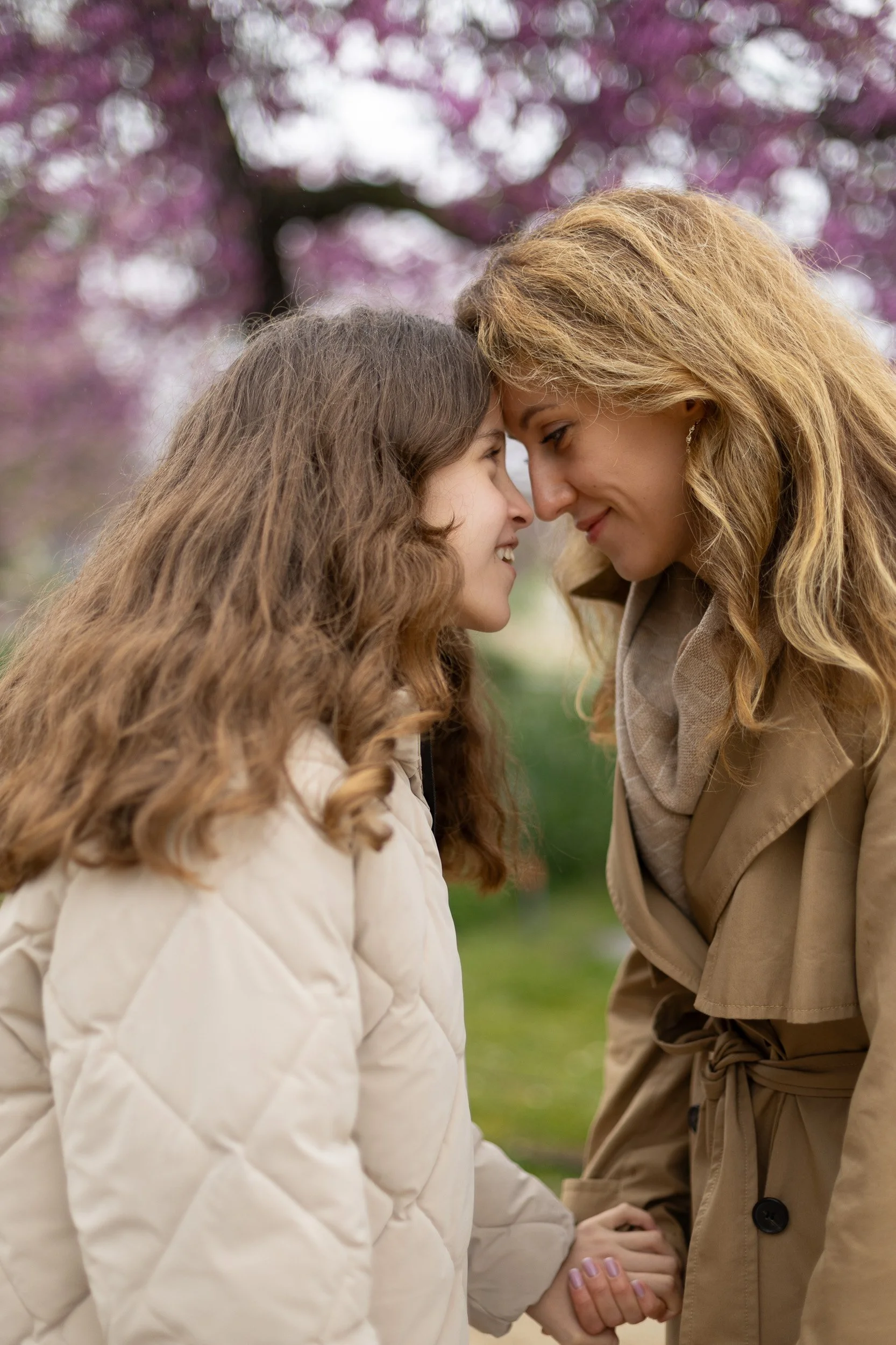A woman and a girl are touching foreheads and smiling at each other outdoors during spring with pink blossoms in the background.