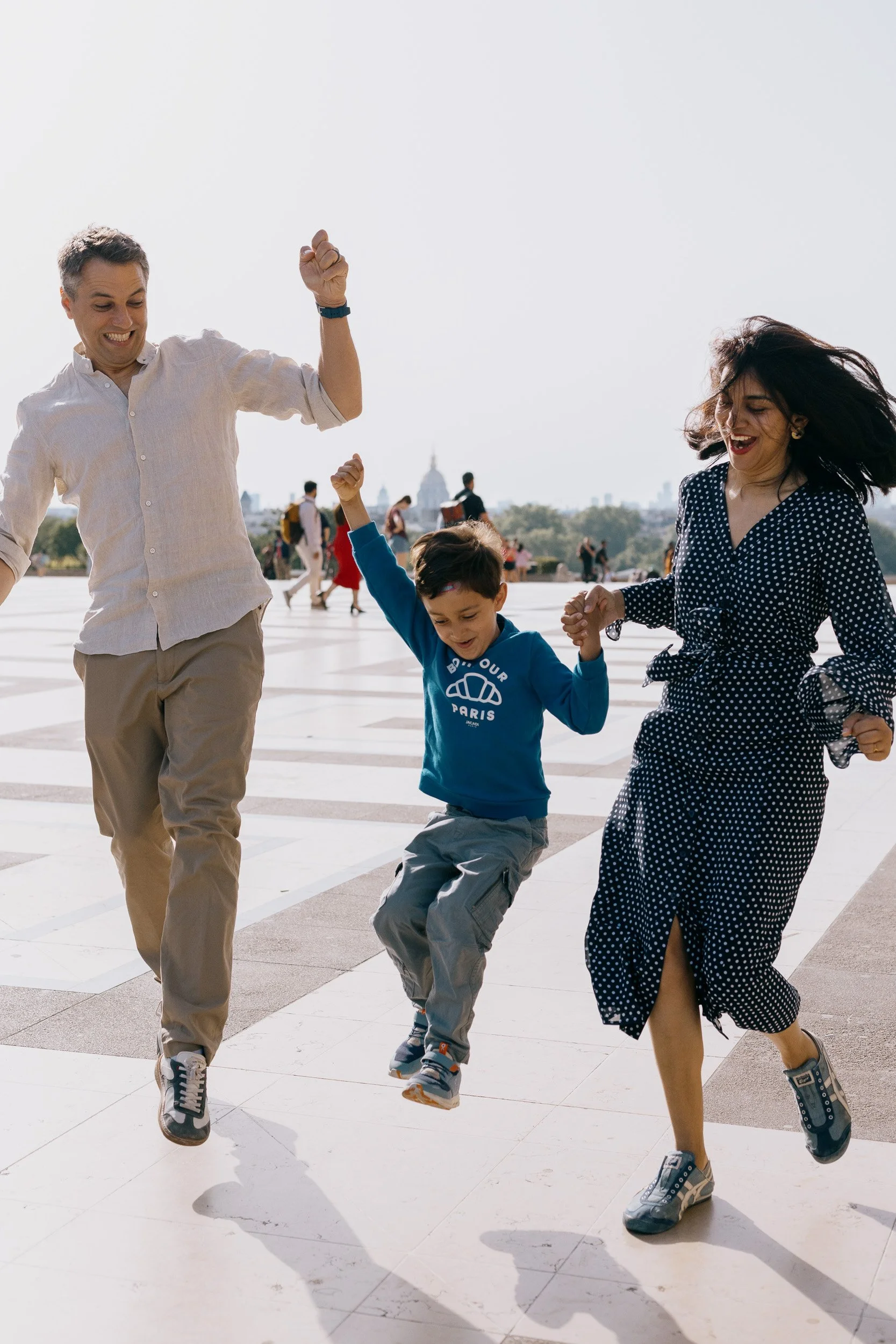 A family of three jumping and holding hands outdoors on a sunny day, with a city skyline in the background.