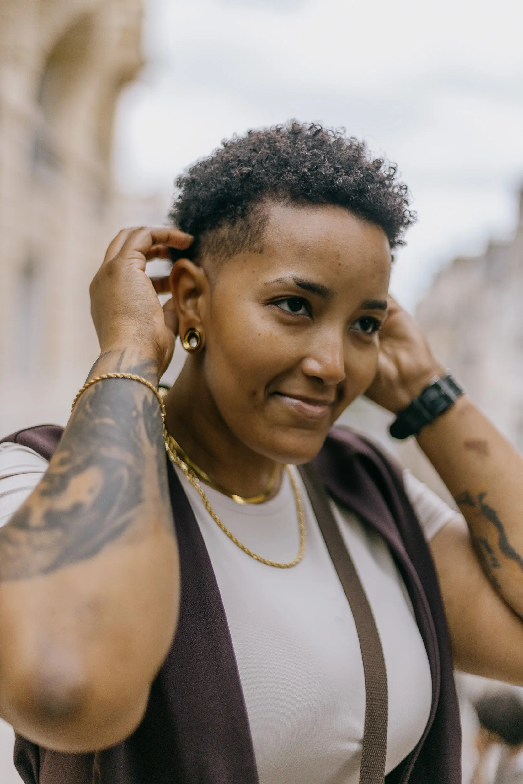 Close-up of a woman with short curly hair, wearing earrings and a gold chain, adjusting her hair with a slight smile, outdoors.