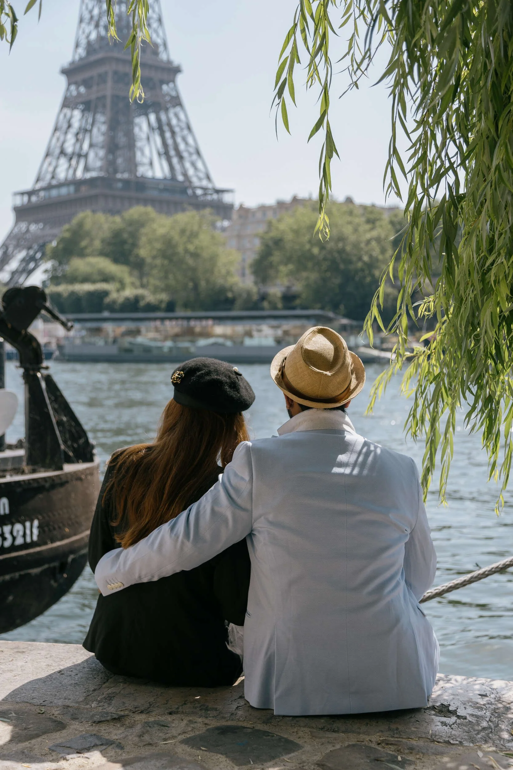 A couple sitting close together on a stone bench by the river, with the Eiffel Tower partially obscured behind trees in Paris, France. The man is wearing a white jacket and a straw hat, and the woman has long hair and is wearing a black jacket.