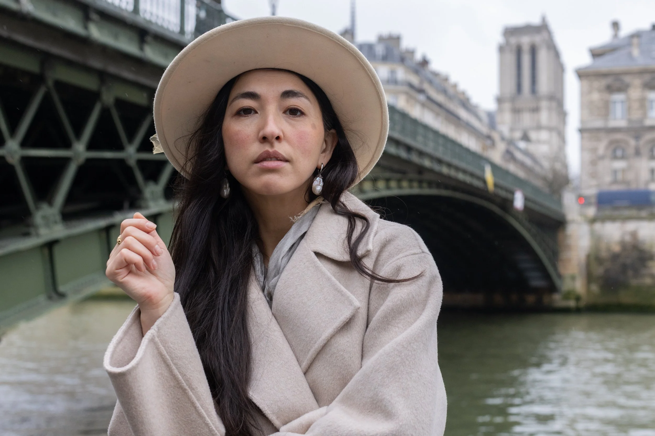 A woman wearing a beige hat and coat standing under a bridge in an urban area with historic buildings in the background.