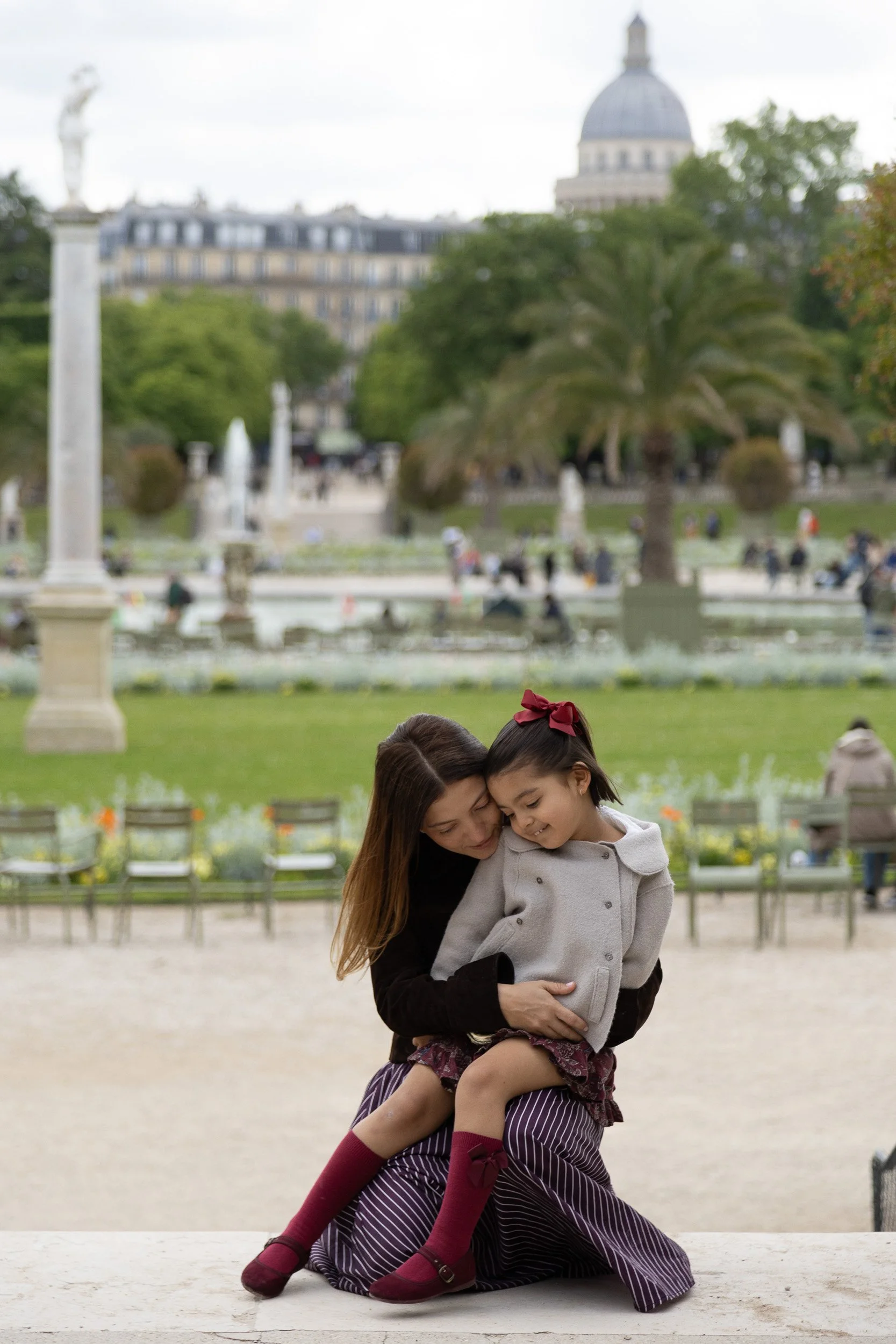 A woman hugging a young girl in a park with a fountain, trees, and historic buildings in the background.