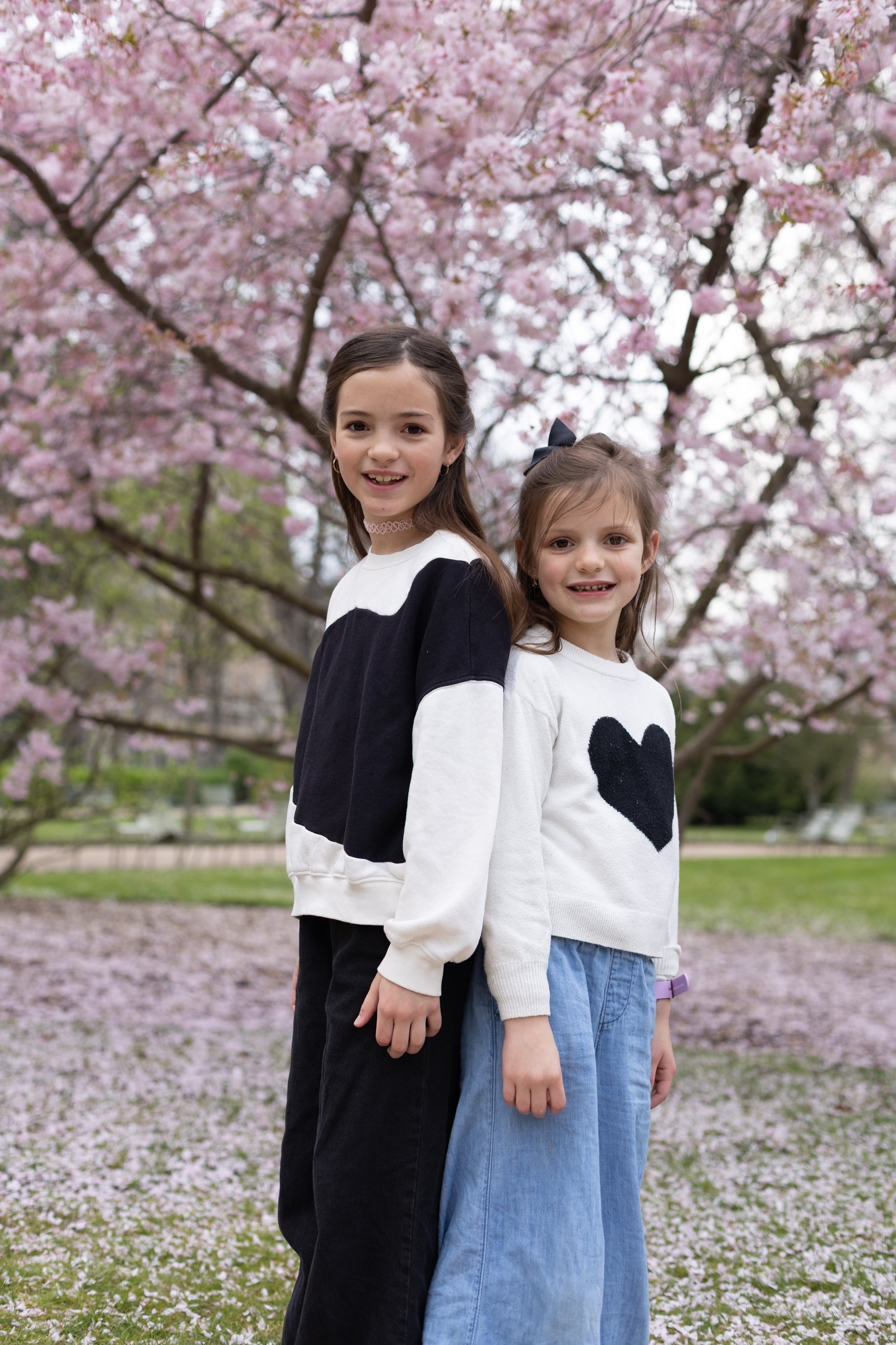 Two young girls standing back to back outdoors in a park with pink cherry blossom trees in the background.