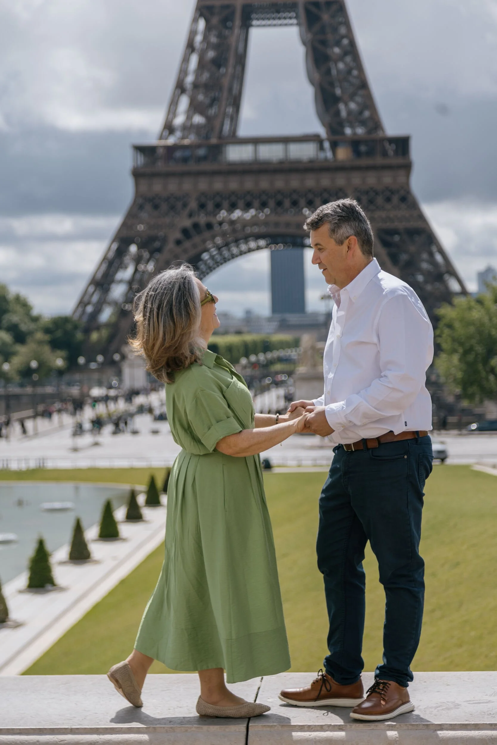 A couple holding hands and looking at each other near the Eiffel Tower in Paris, France, with the woman wearing a green dress and sunglasses and the man wearing a white shirt and dark pants, both smiling, on a cloudy day.