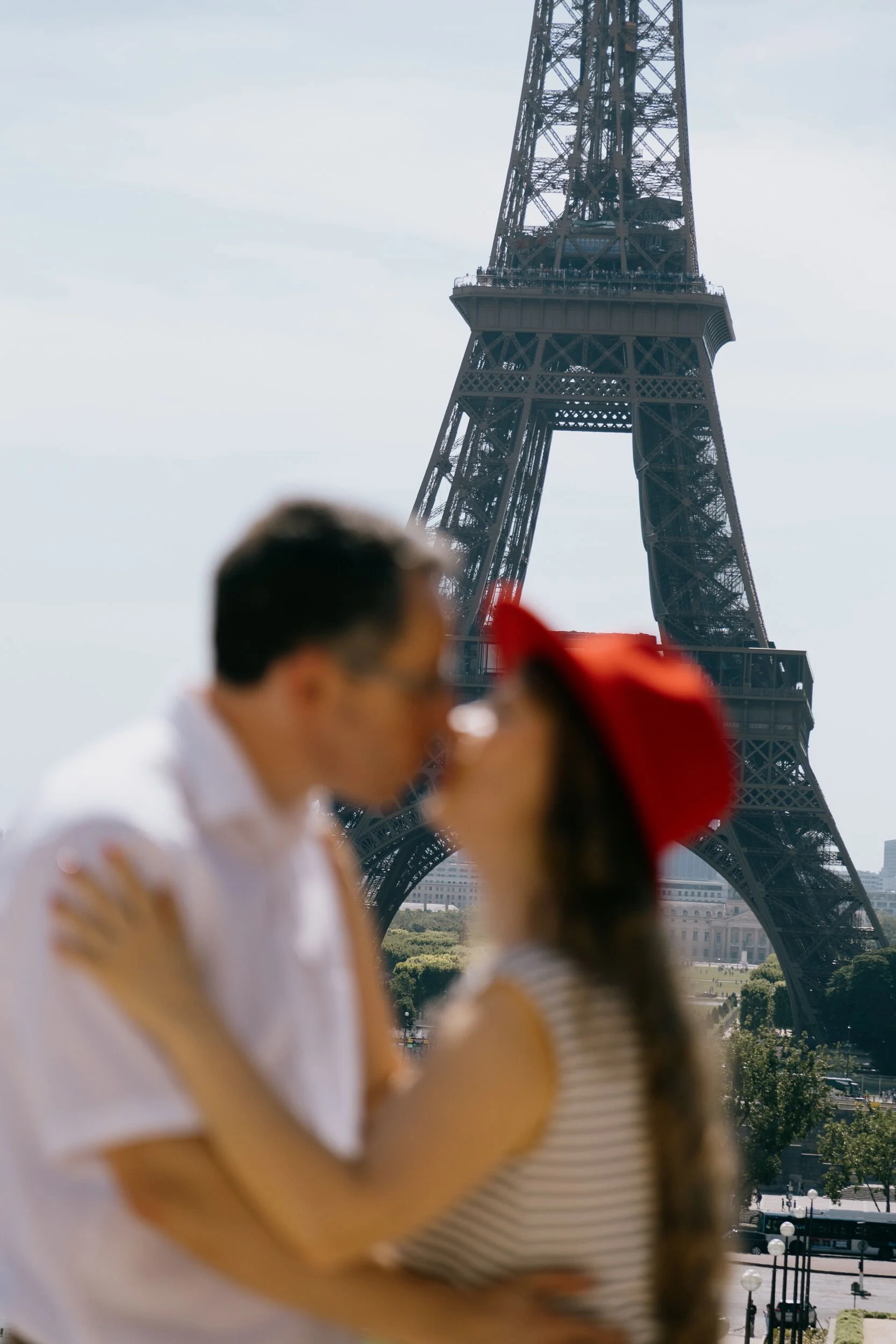 A couple is standing near the Eiffel Tower in Paris, with a cityscape and greenery in the background. The man is wearing a white shirt, and the woman is wearing a black and white striped shirt and a red hat.