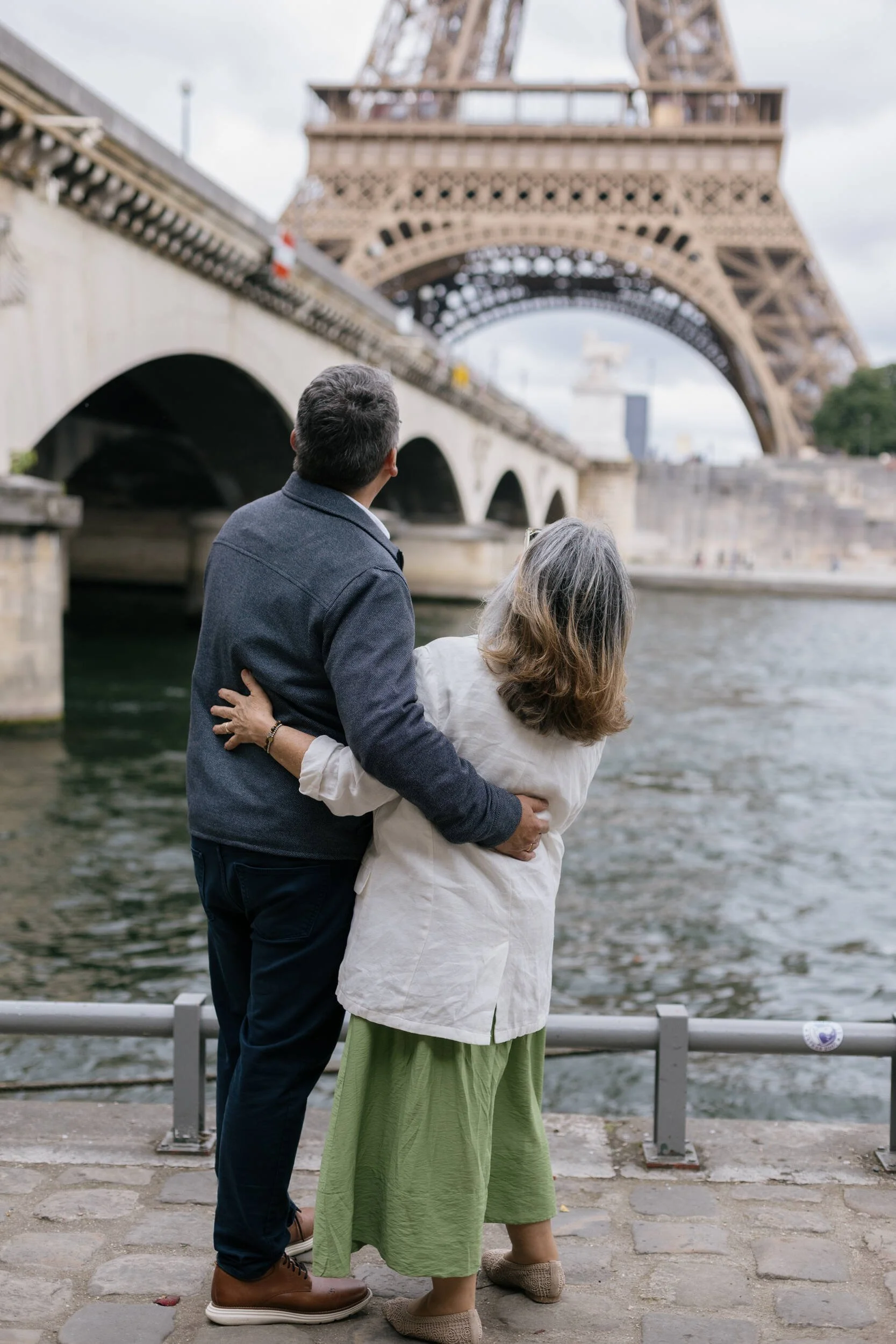 A man and a woman stand close together, looking at the Eiffel Tower in Paris, France. The man has his arm around the woman's shoulder, and they are looking up at the iconic iron tower.