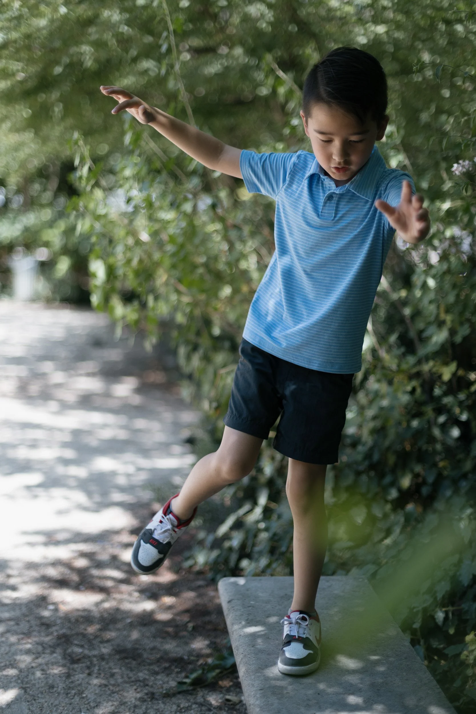 Young boy in a blue striped shirt and shorts balancing on a concrete bench, surrounded by greenery.