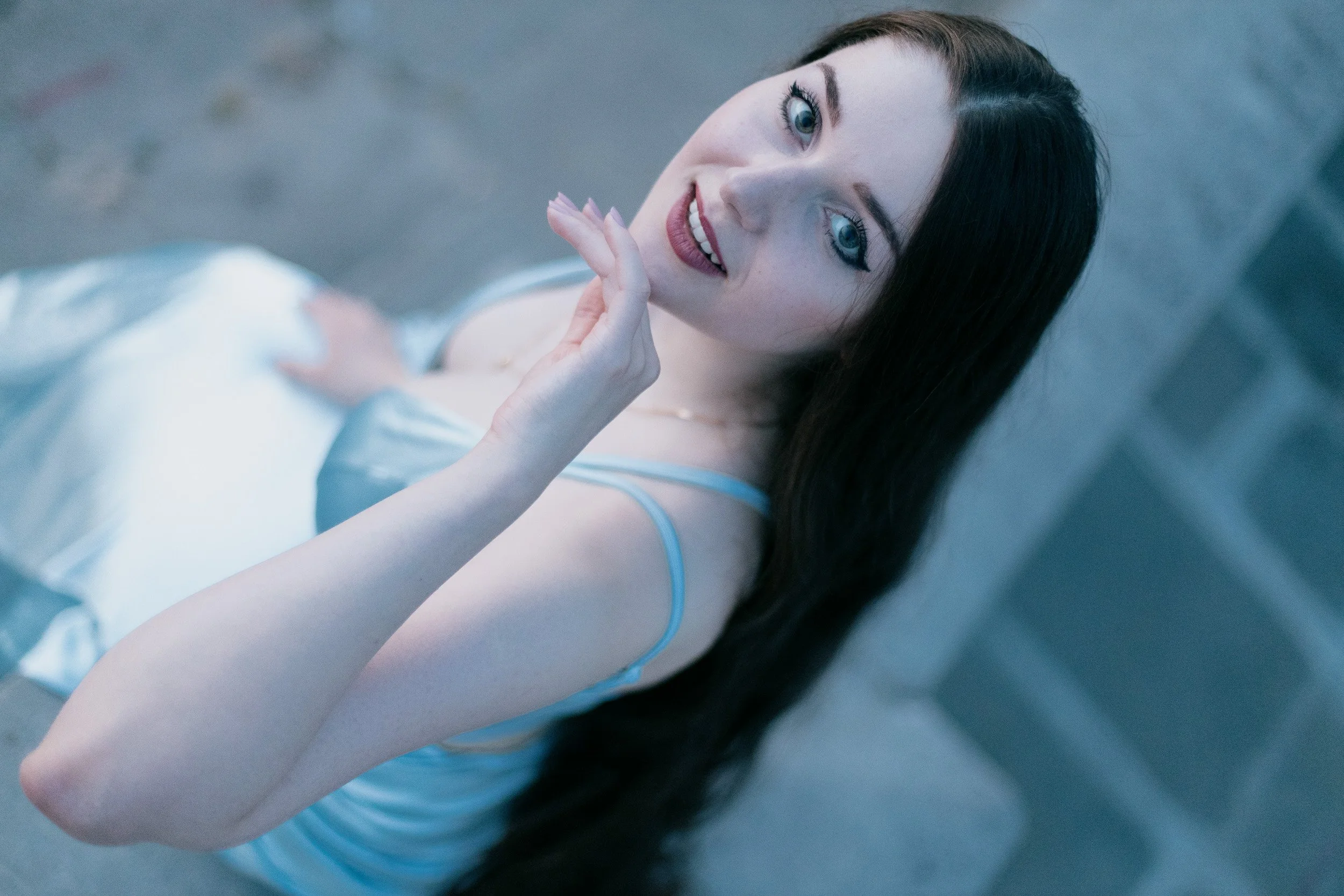 Woman with black hair and blue eyes lying down on a grey surface, smiling and holding her chin with her left hand, wearing a white satin top.