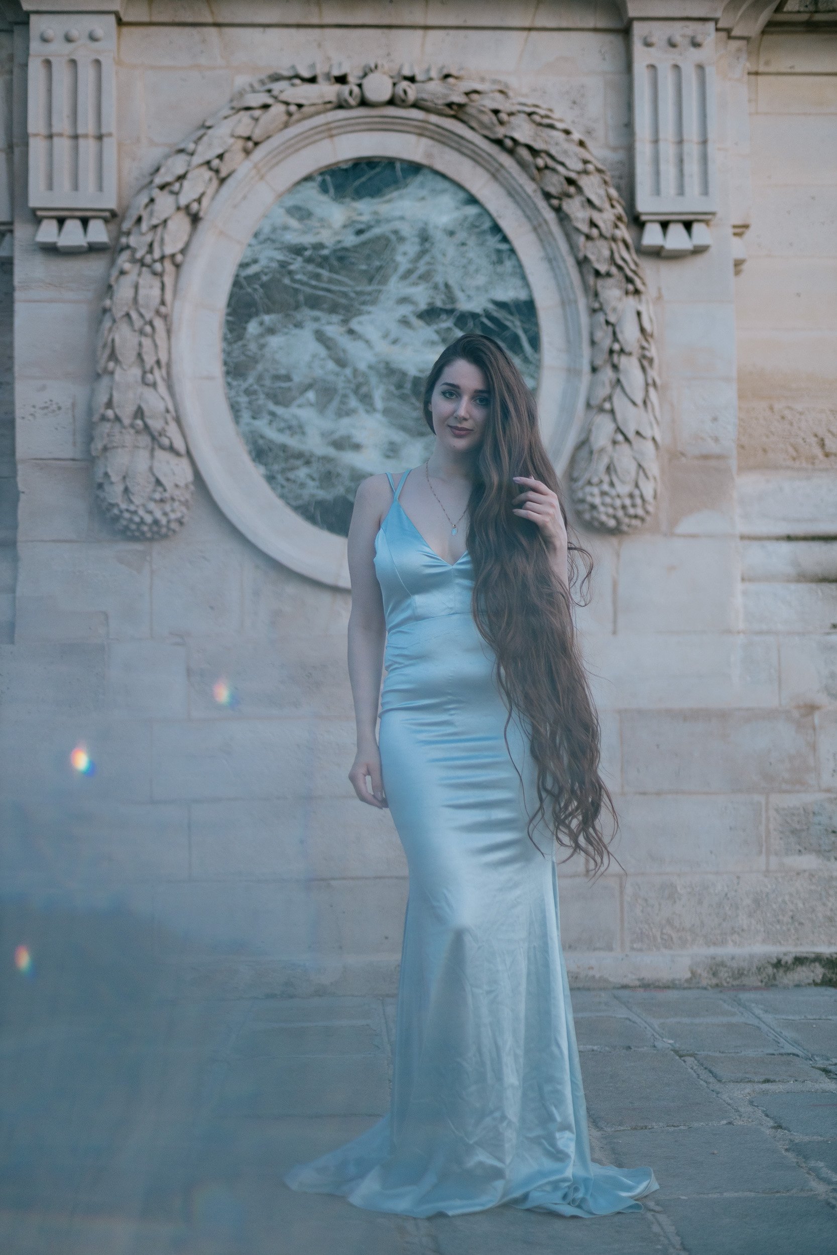 A woman with long, wavy brown hair wearing a light blue satin gown standing in front of a stone wall with an ornate mirror reflecting a body of water.