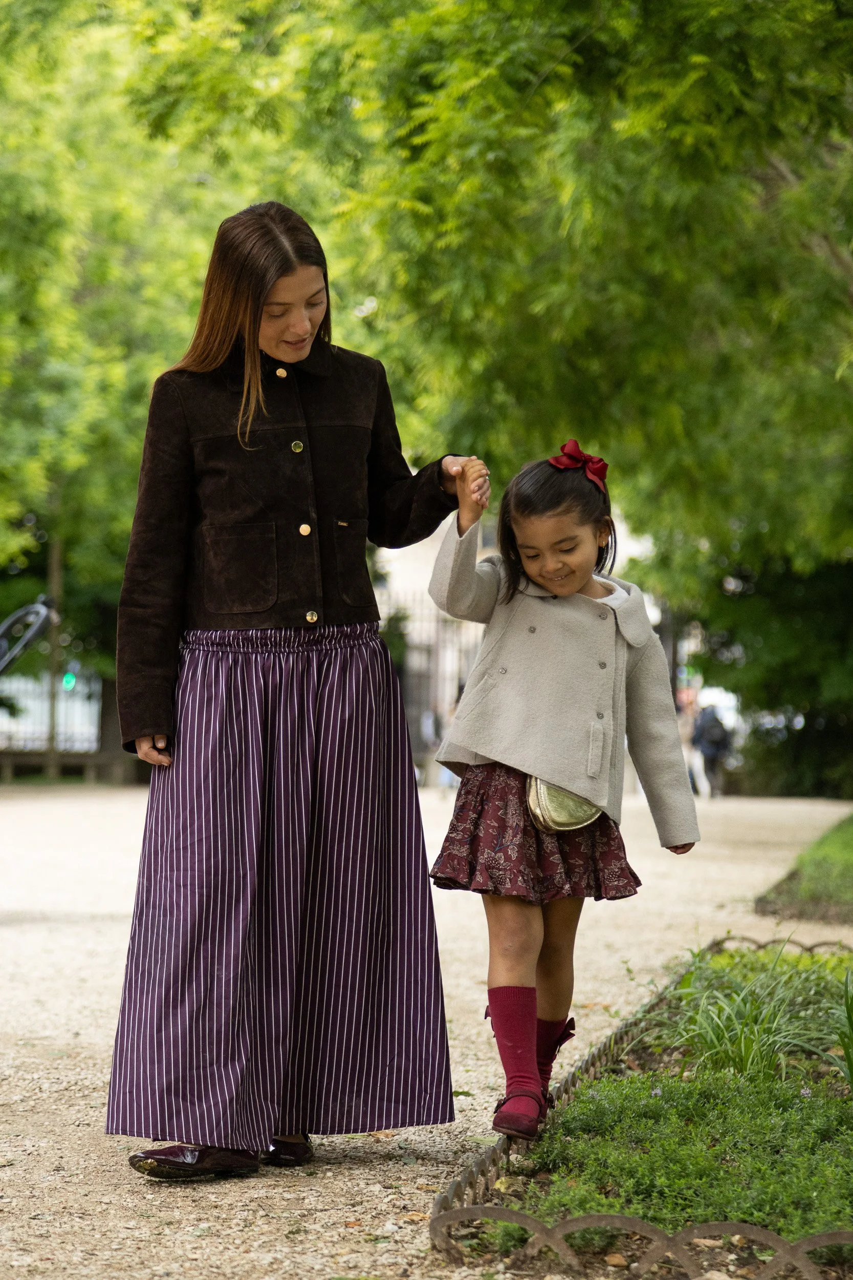 A woman and young girl walk hand-in-hand in a park surrounded by green trees. The woman wears a dark jacket and striped wide-leg pants, while the girl wears a beige coat, red skirt, and maroon boots with a red bow in her hair.