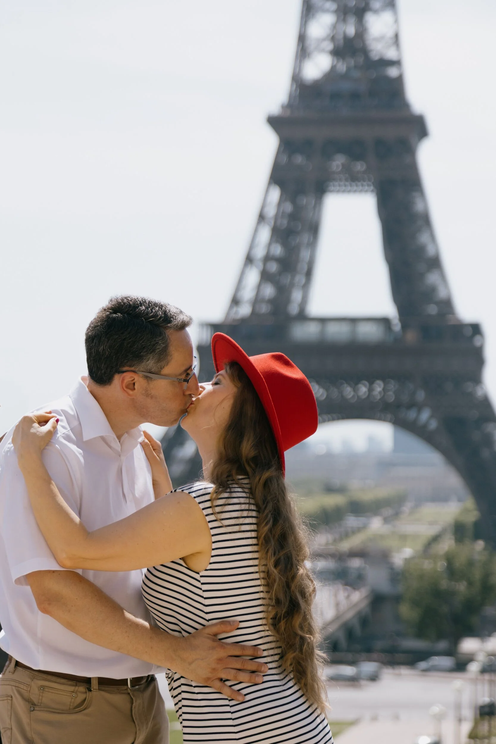 A couple kissing in front of the Eiffel Tower in Paris, France. The woman is wearing a striped dress and a red hat, and the man is wearing a white shirt and glasses.