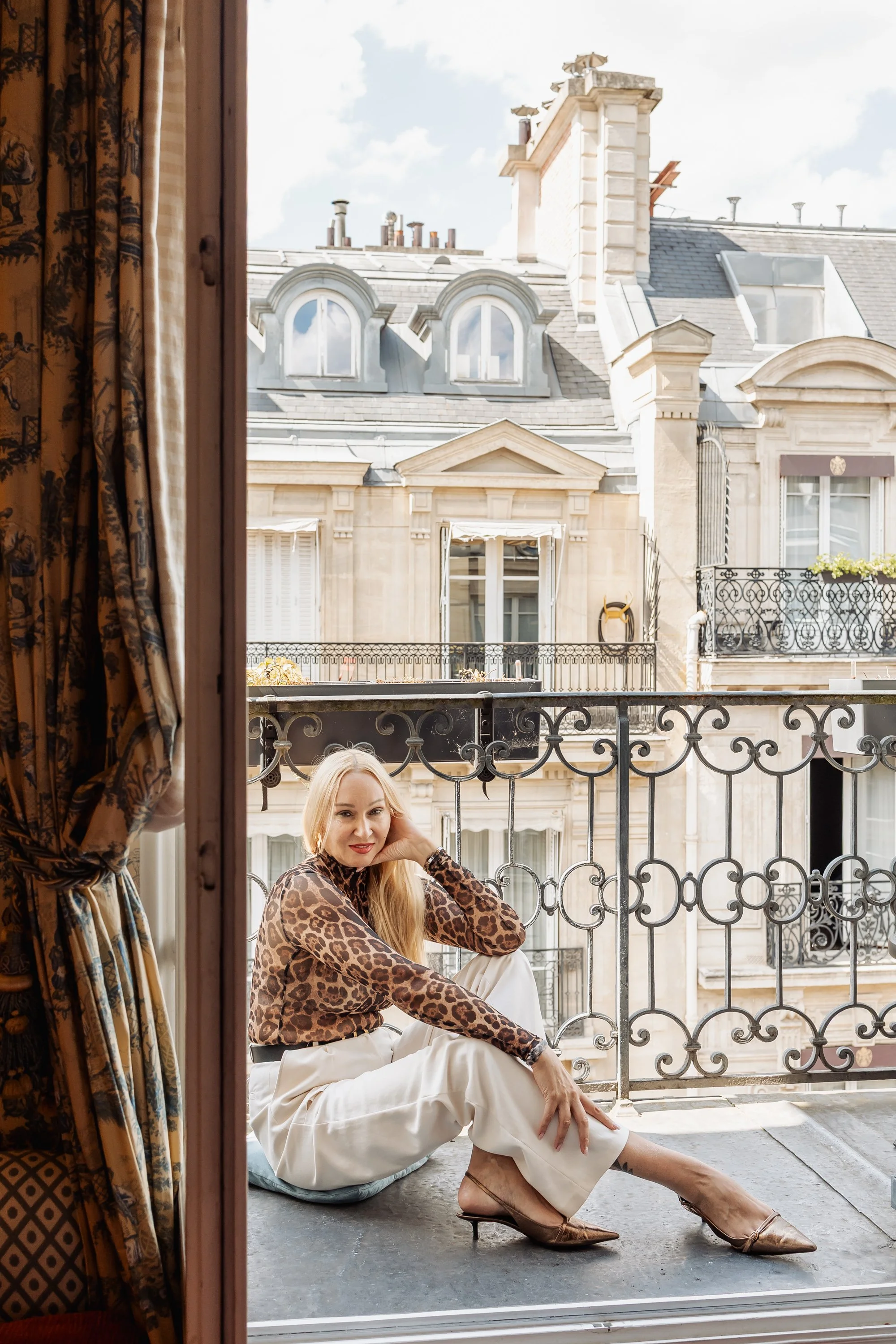 A woman with blonde hair, wearing a leopard print top and white pants, sitting on a balcony with a decorative metal railing, with buildings and a partly cloudy sky in the background.